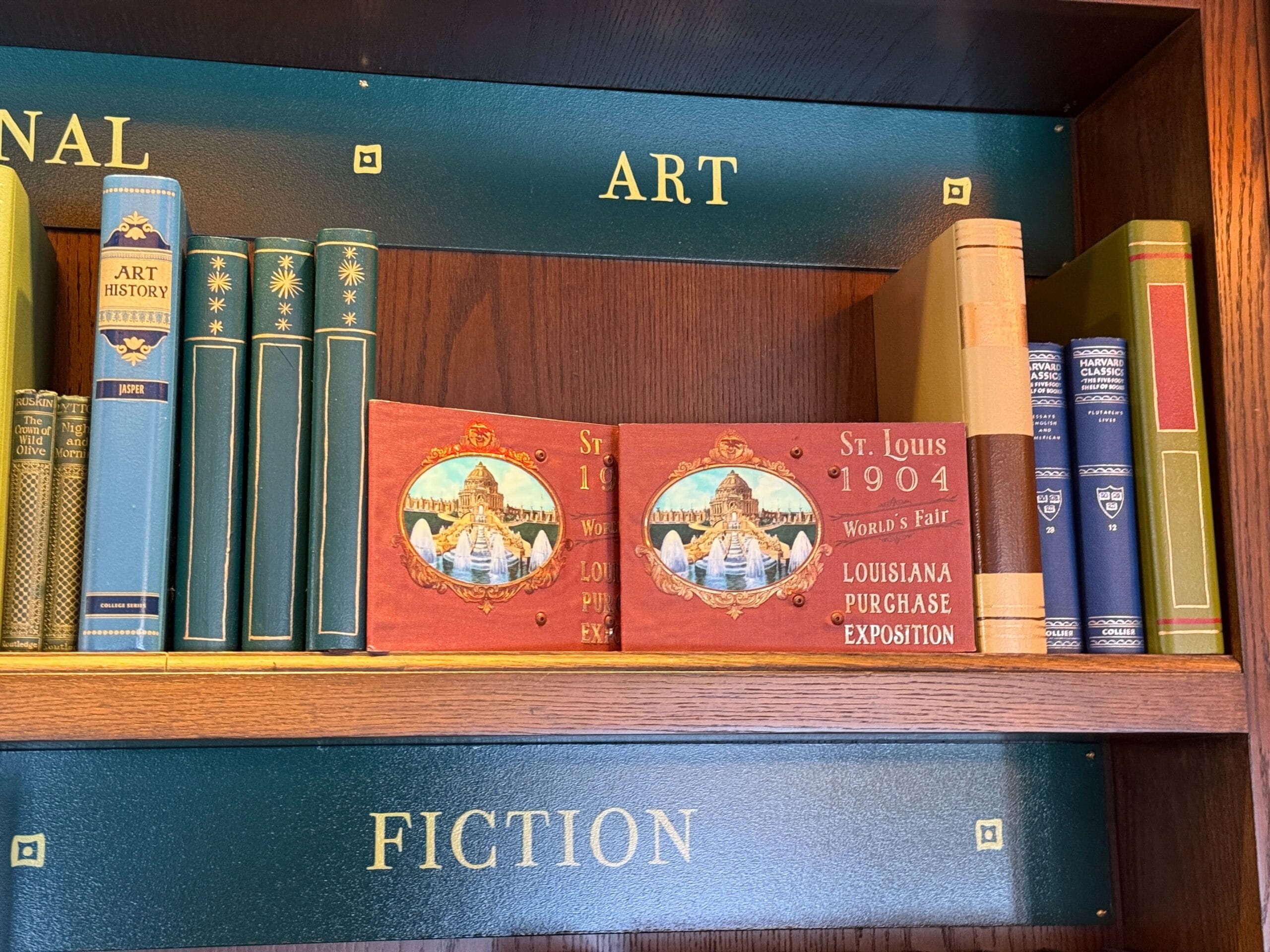 Bookshelf marked "ART" with blue and green books and two red St. Louis World’s Fair souvenir books, reminiscent of Disney park displays.