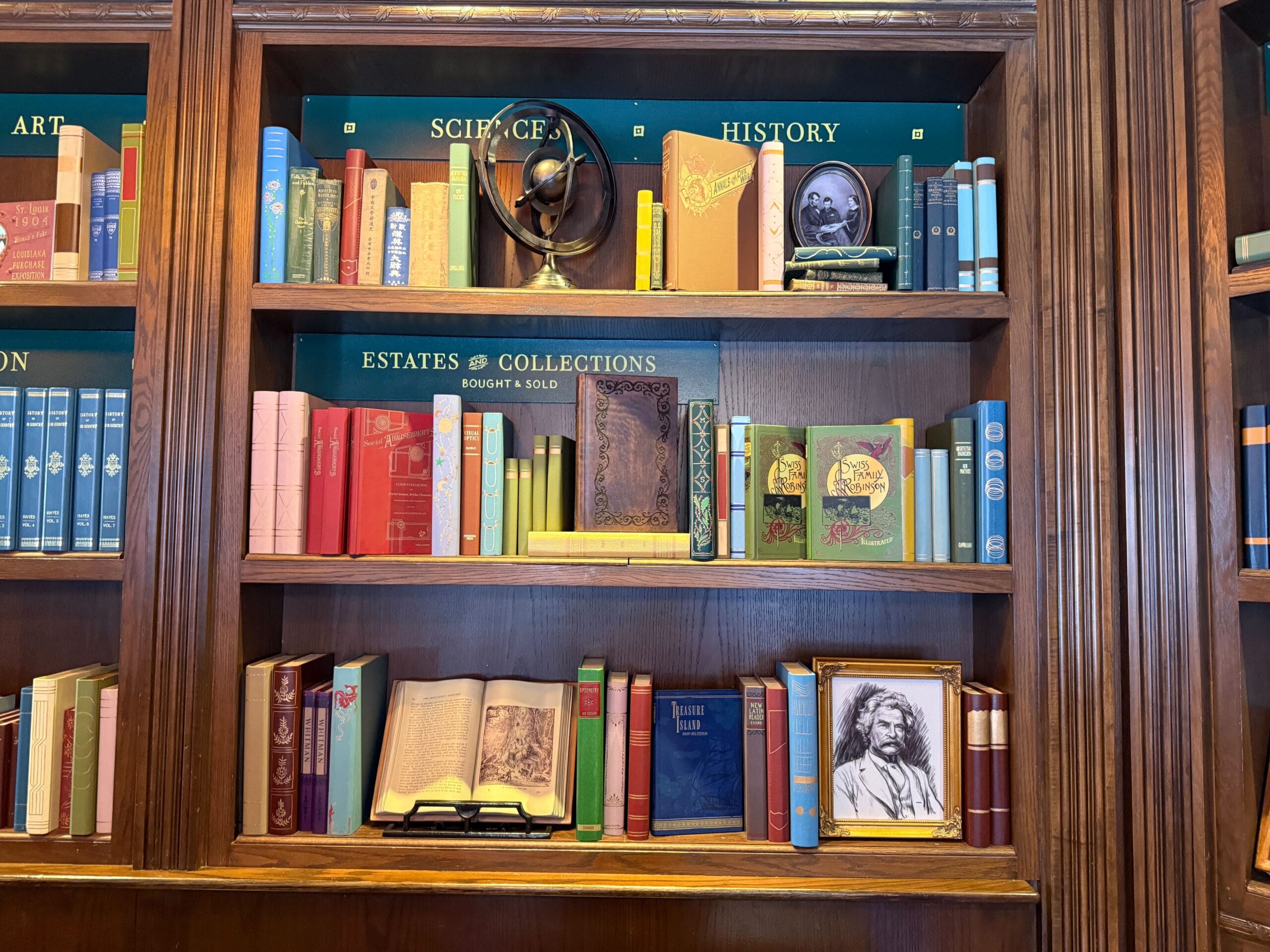 Bookshelves filled with colorful books, Disney park memorabilia, and framed photos under science, history, and estate signs.