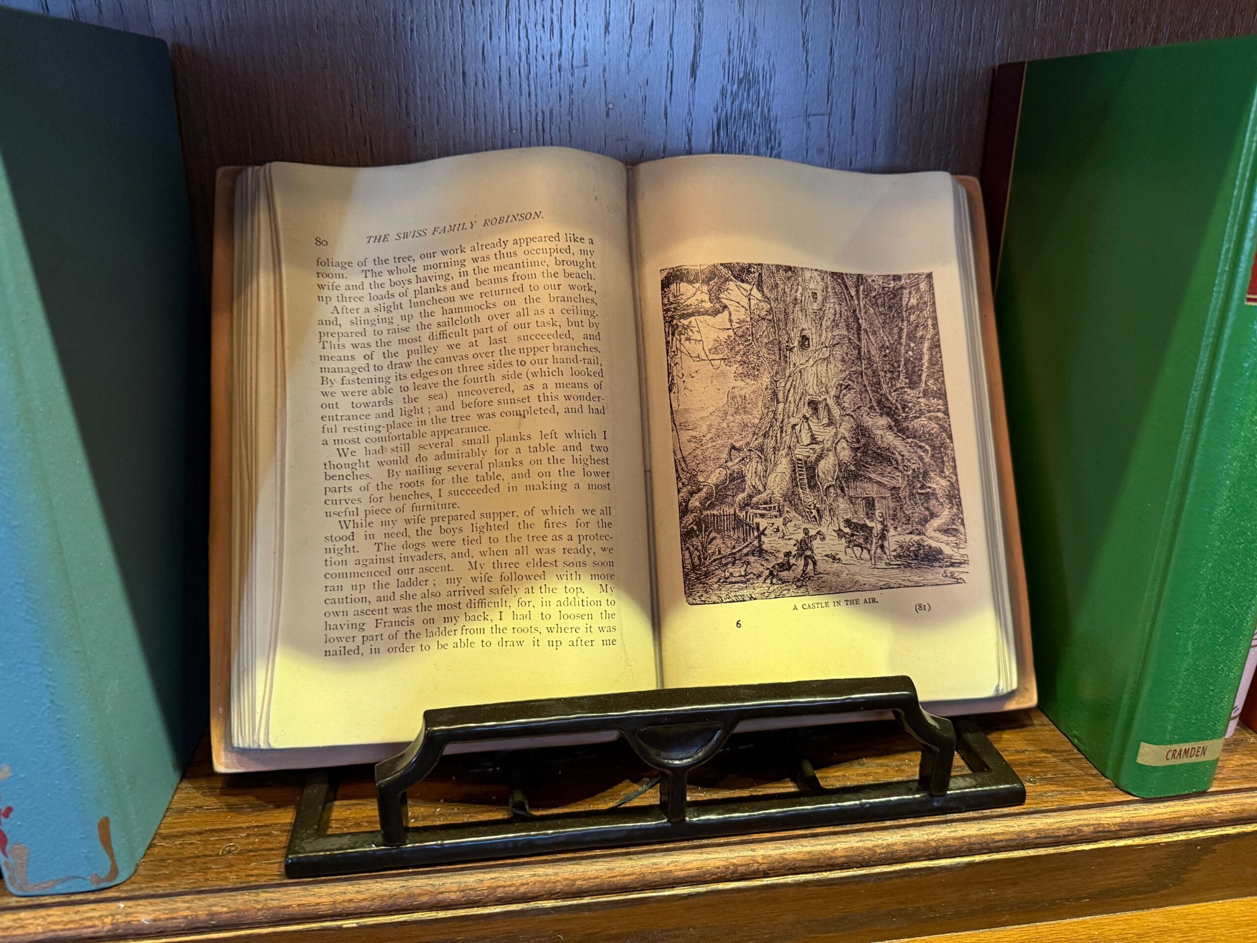 An open book with text and a black-and-white illustration on a wooden shelf, flanked by closed books, like in a Disney park library.
