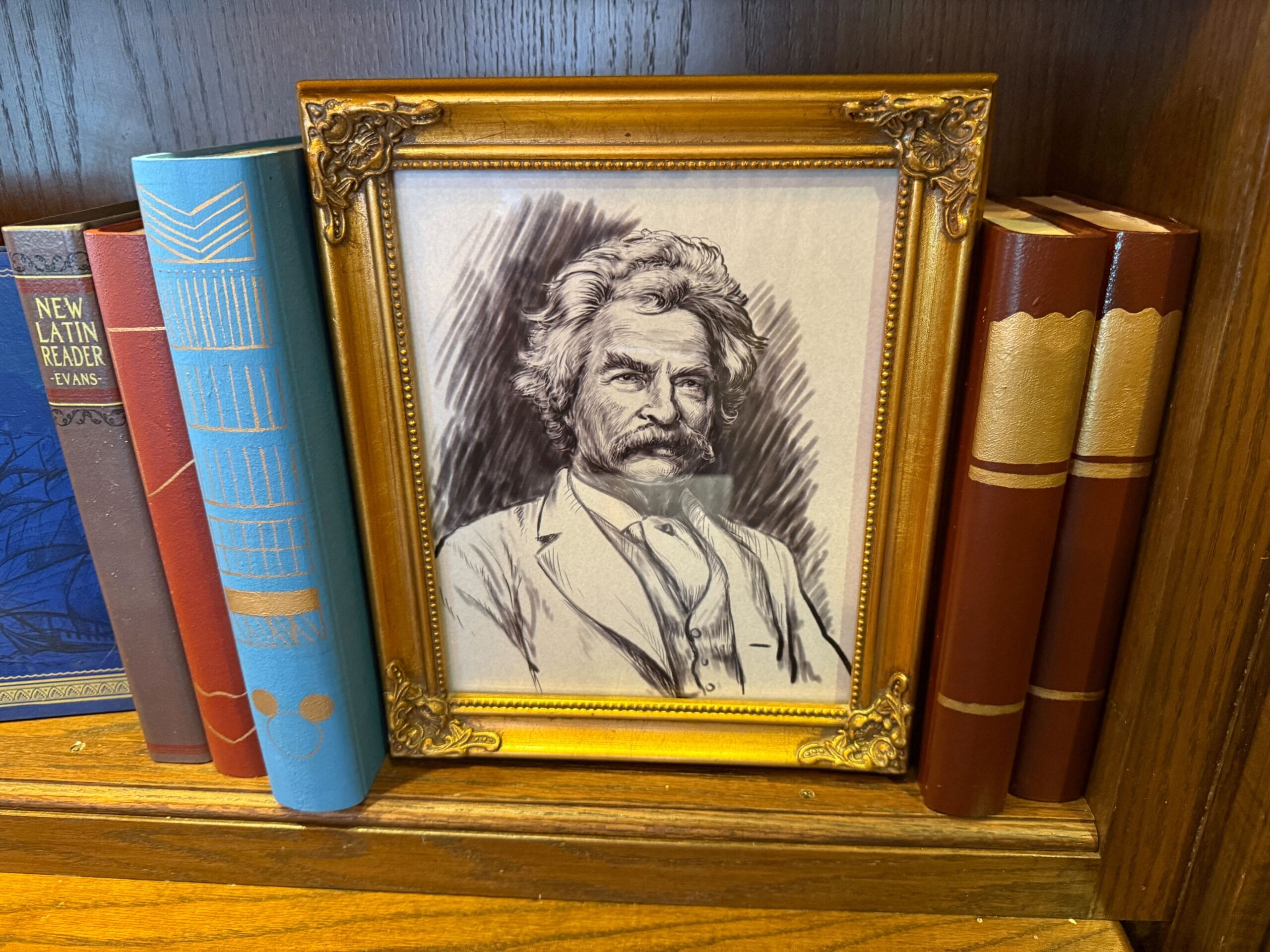 A framed black-and-white portrait of a mustached man sits on a bookshelf among classic books, reminiscent of Walt Disney’s study.