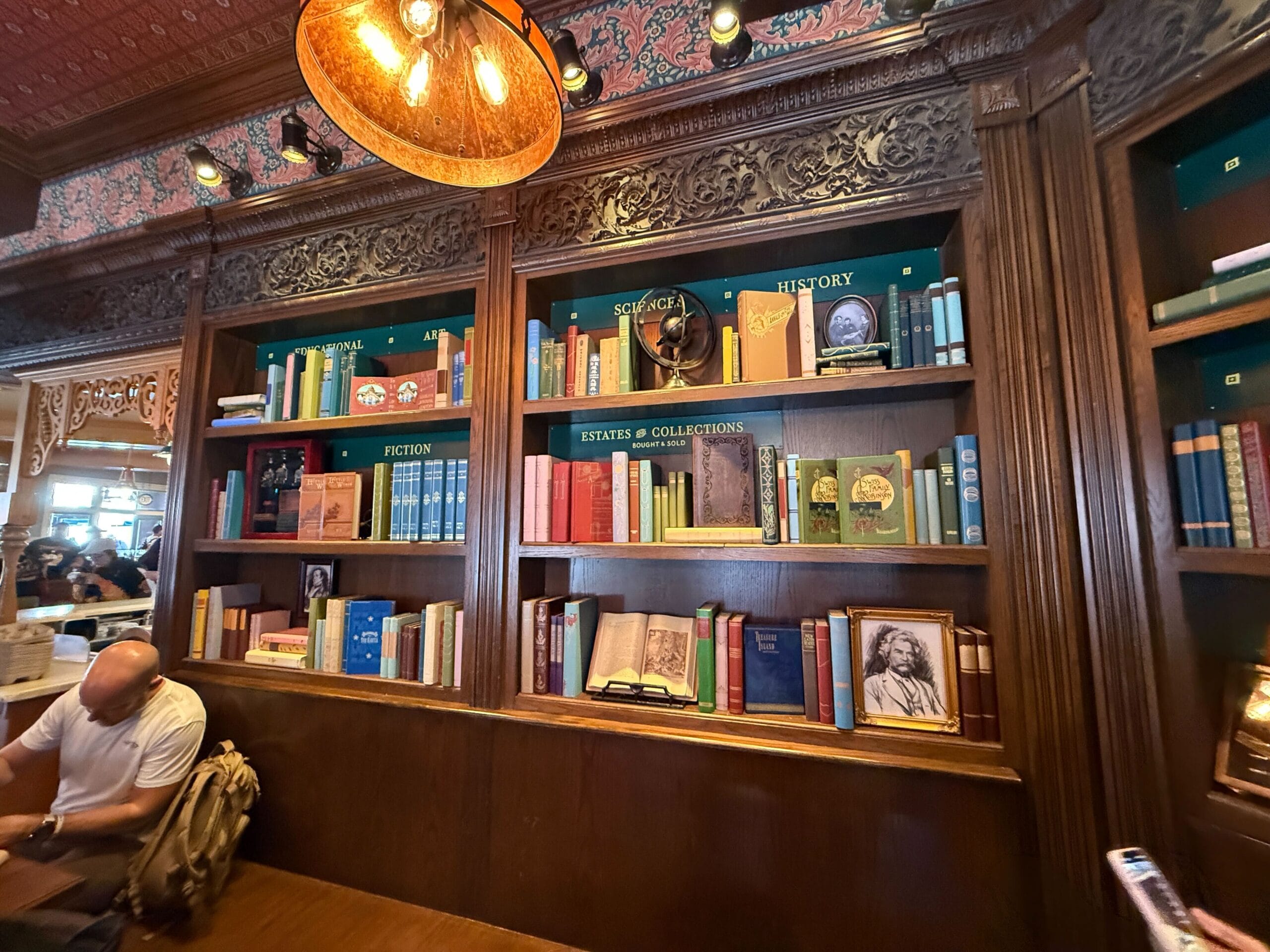 A wooden bookshelf with colorful books and a man reading in a cozy room, reminiscent of Disney’s library-themed park interiors.