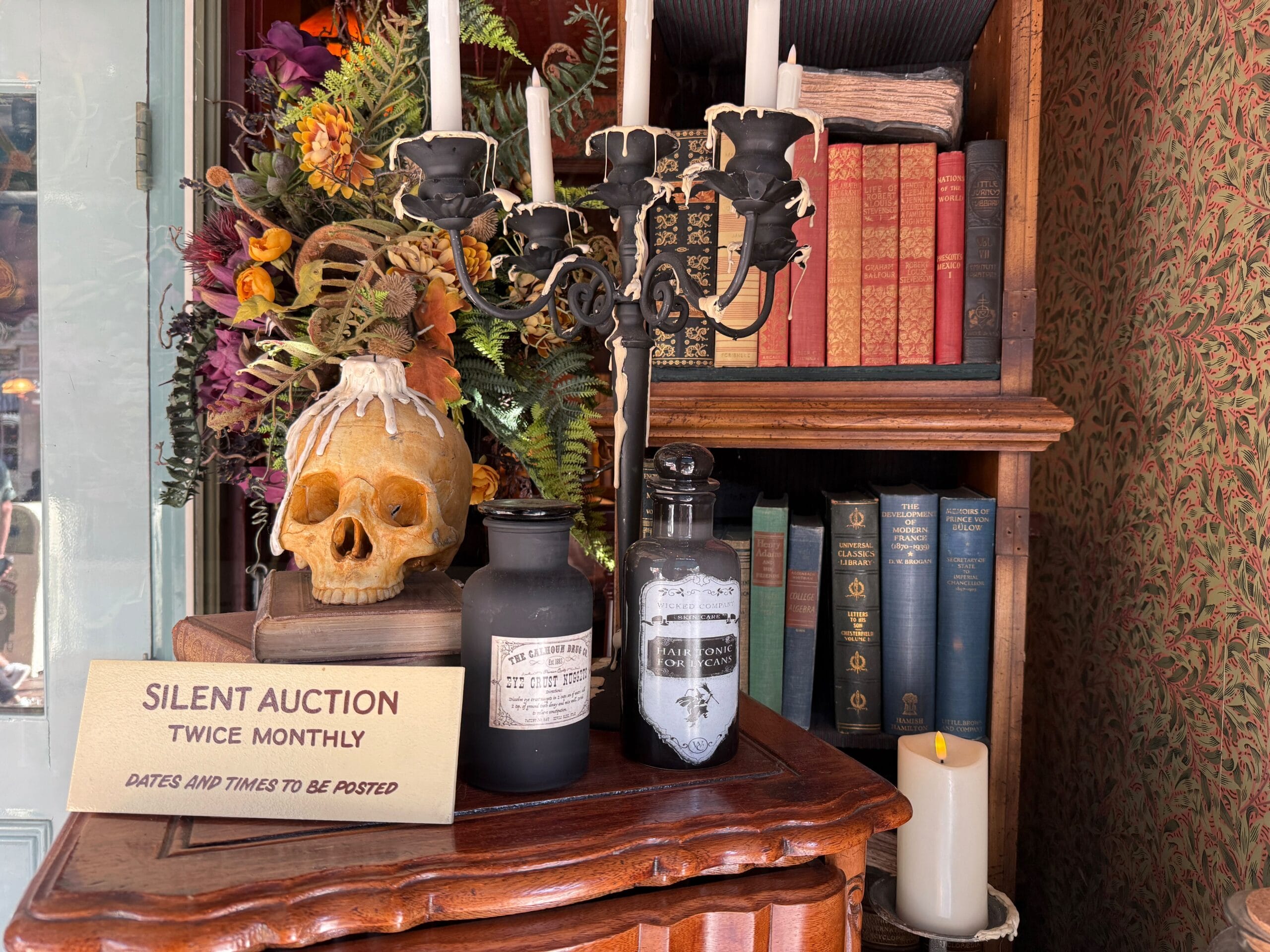 A skull, candles, and old books on a wooden shelf evoke Haunted Mansion vibes, with a “Silent Auction Twice Monthly” sign in front.