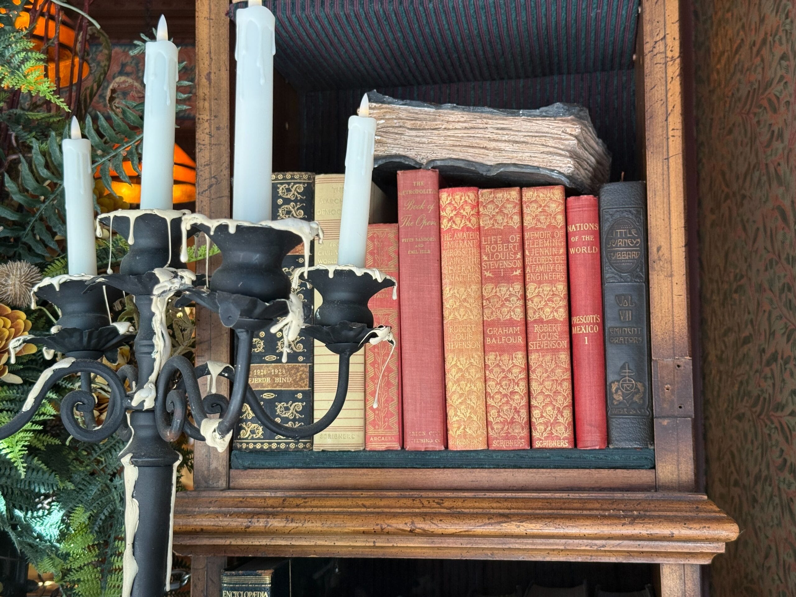 A black candle holder with white candles sits before vintage books on a wooden shelf, evoking the classic library vibe of Disney parks.