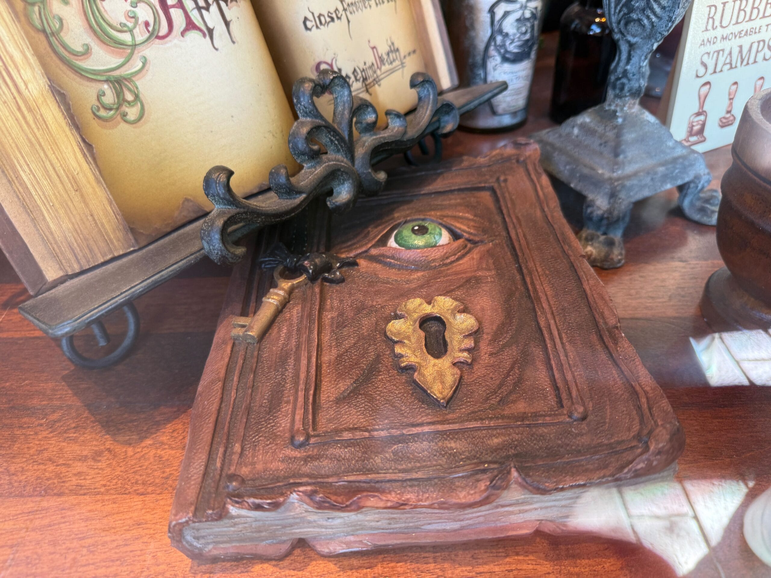 A brown book with a sculpted green eye and keyhole on the cover rests on wood, evoking the mysterious artifacts found at Disney parks.