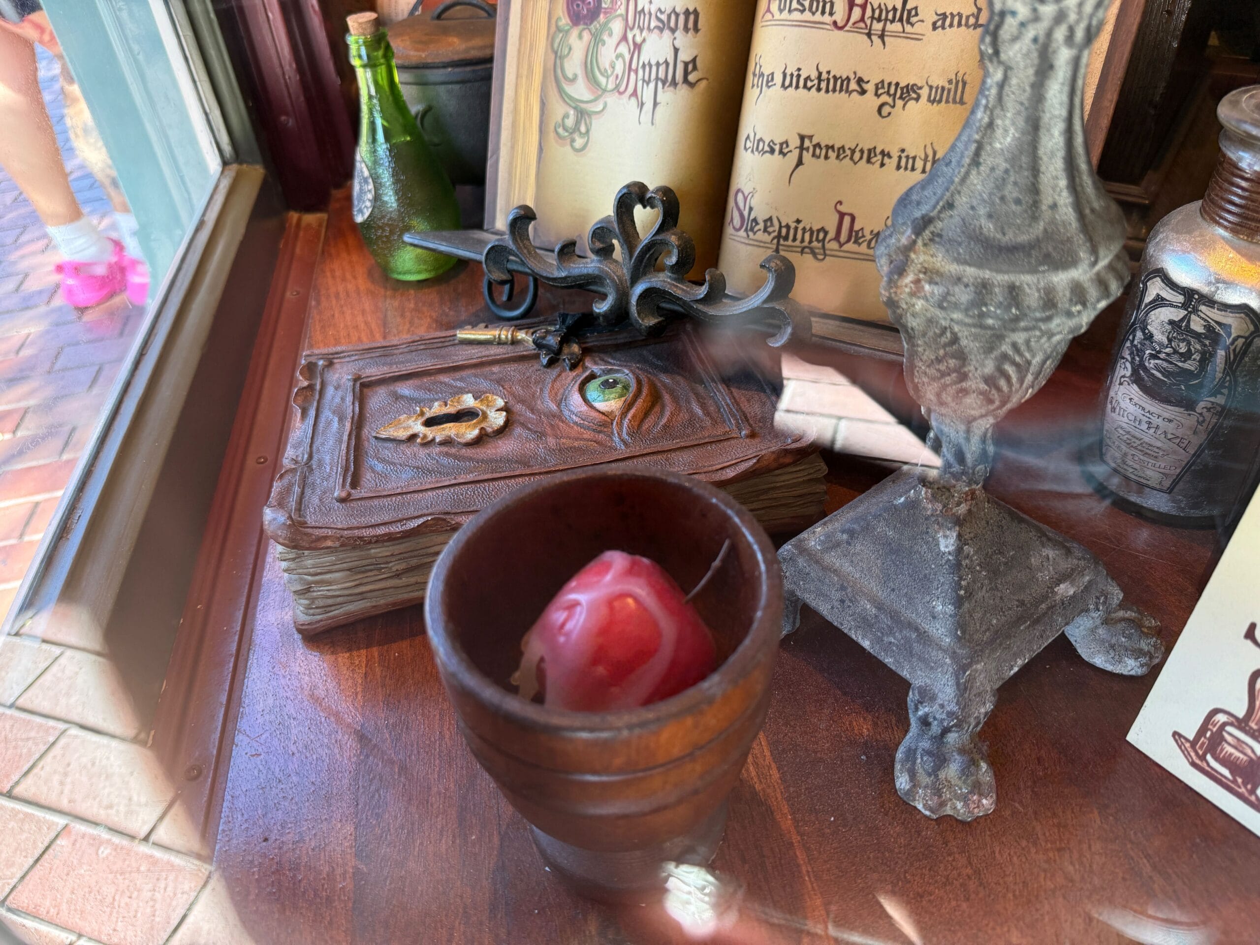 On a wooden table at Disney park, a red candle, mystical book with an eye, potions, and metal stand evoke magical castle decor.