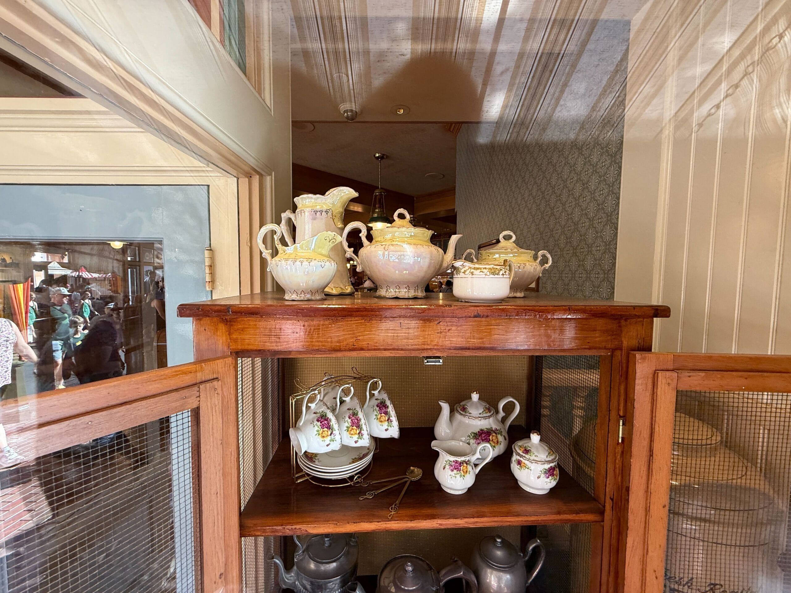 A wooden cabinet at a Disney park displays porcelain teapots, cups, and saucers with floral patterns on two shelves.
