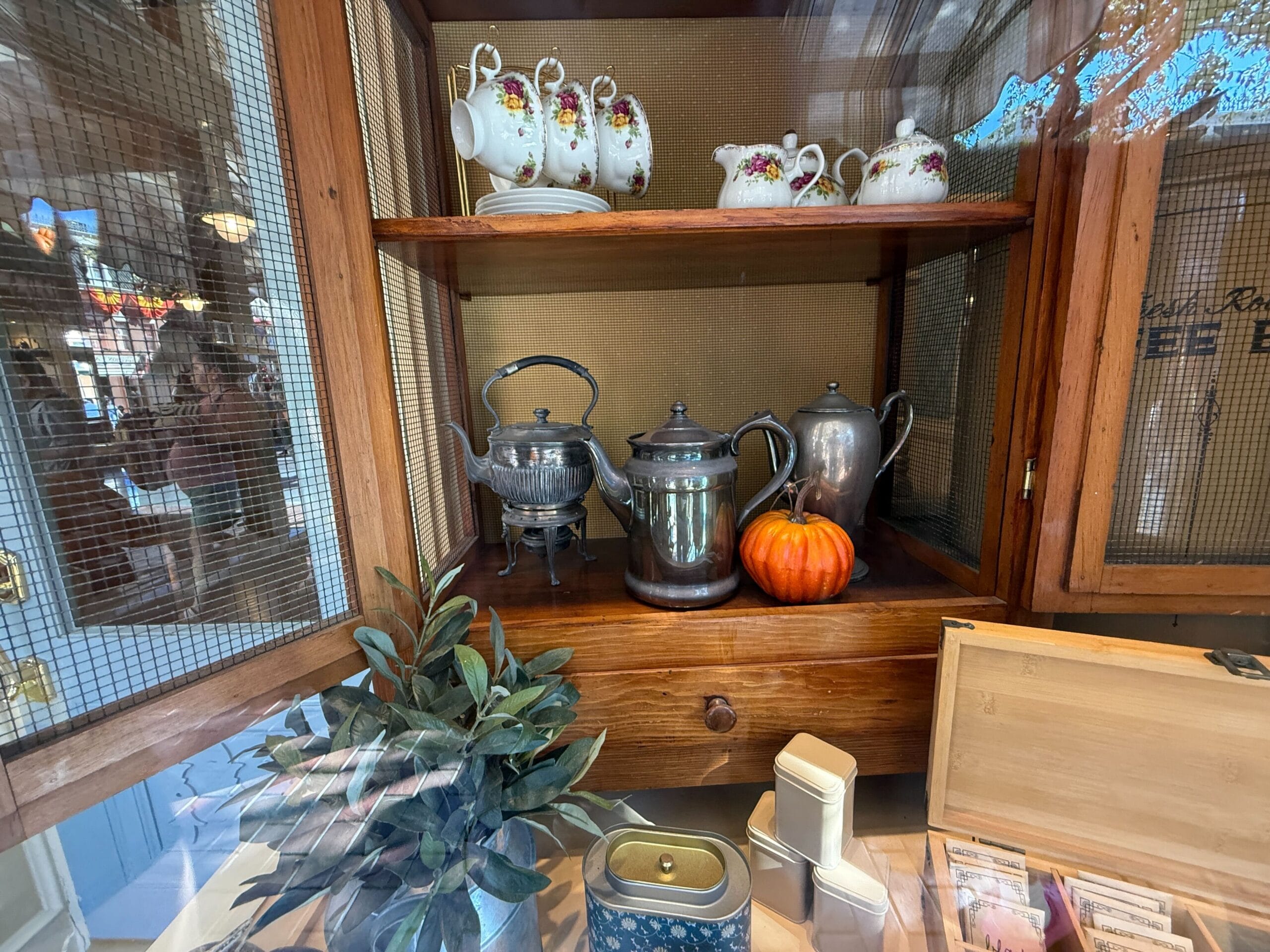 A wooden cabinet, reminiscent of rustic Disney park kitchens, holds teapots, cups, a pumpkin, and utensils behind a mesh door.