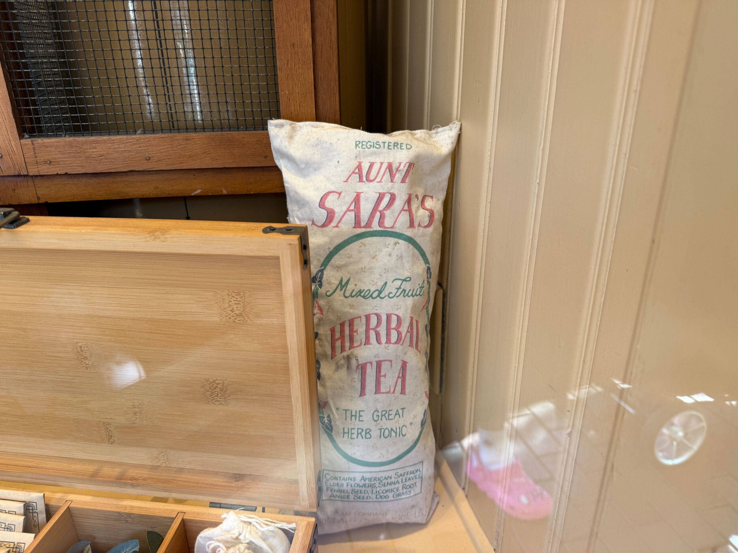 A vintage "Aunt Sara's Herbal Tea" bag, reminiscent of Disney park shop props, stands beside a wooden box on a themed shelf.