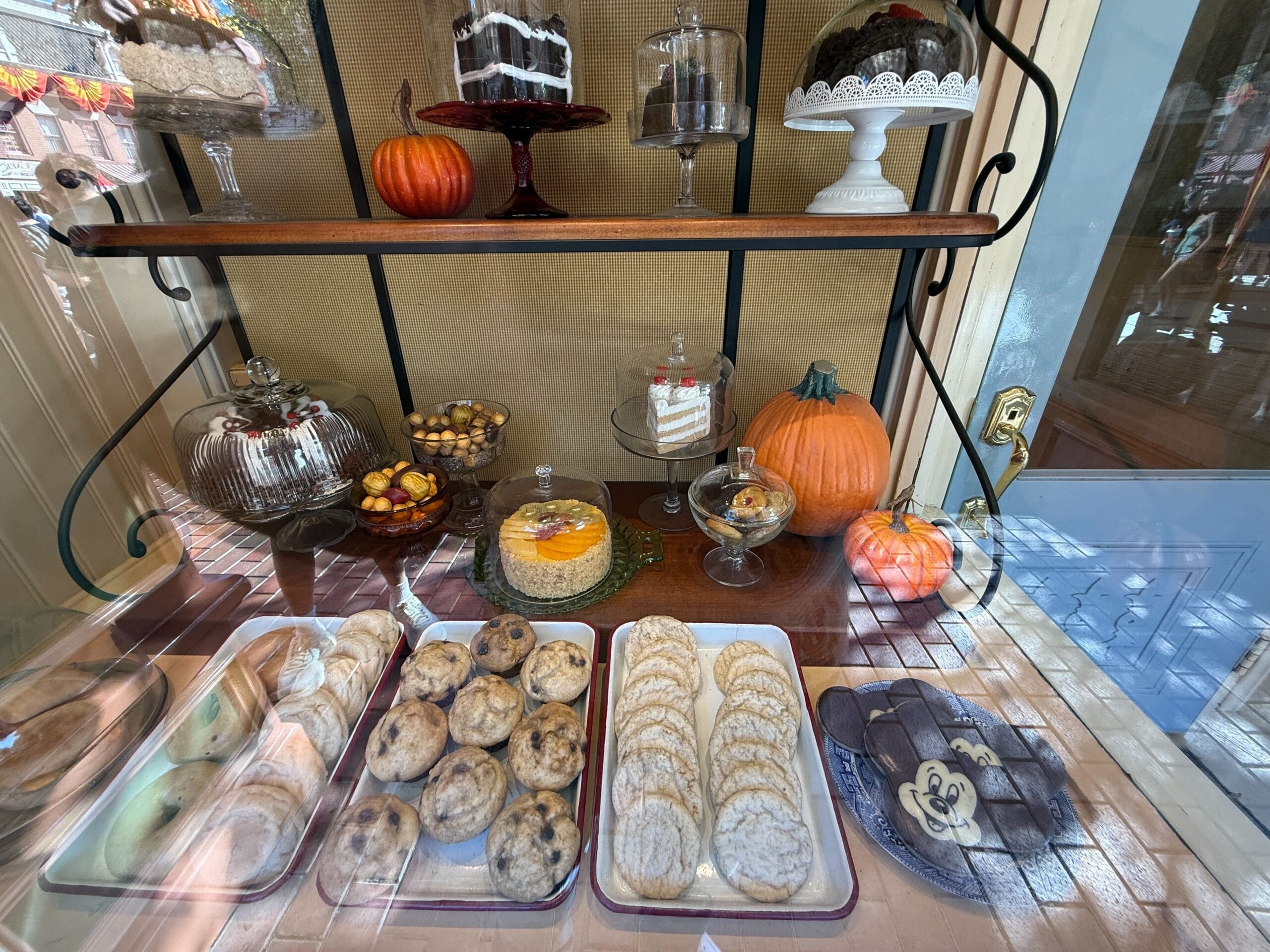 Bakery window at a Disney park with cookies, themed cakes, pumpkins, and desserts on glass stands, evoking Main Street charm.