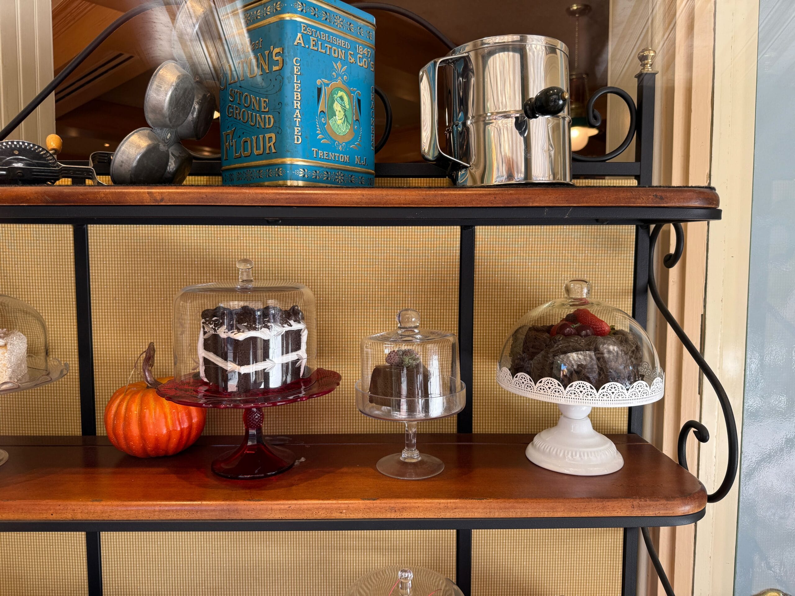 Cakes under glass domes and a pumpkin sit on wooden shelves, with a metal flour tin above—reminiscent of Main Street bakery decor.