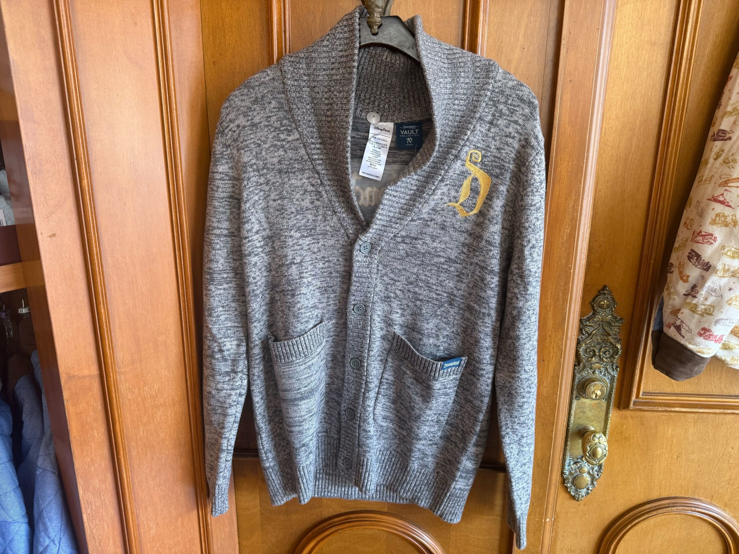 Gray knit cardigan with pockets and yellow "D" on the chest, reminiscent of Disney style, hanging on a wooden door.