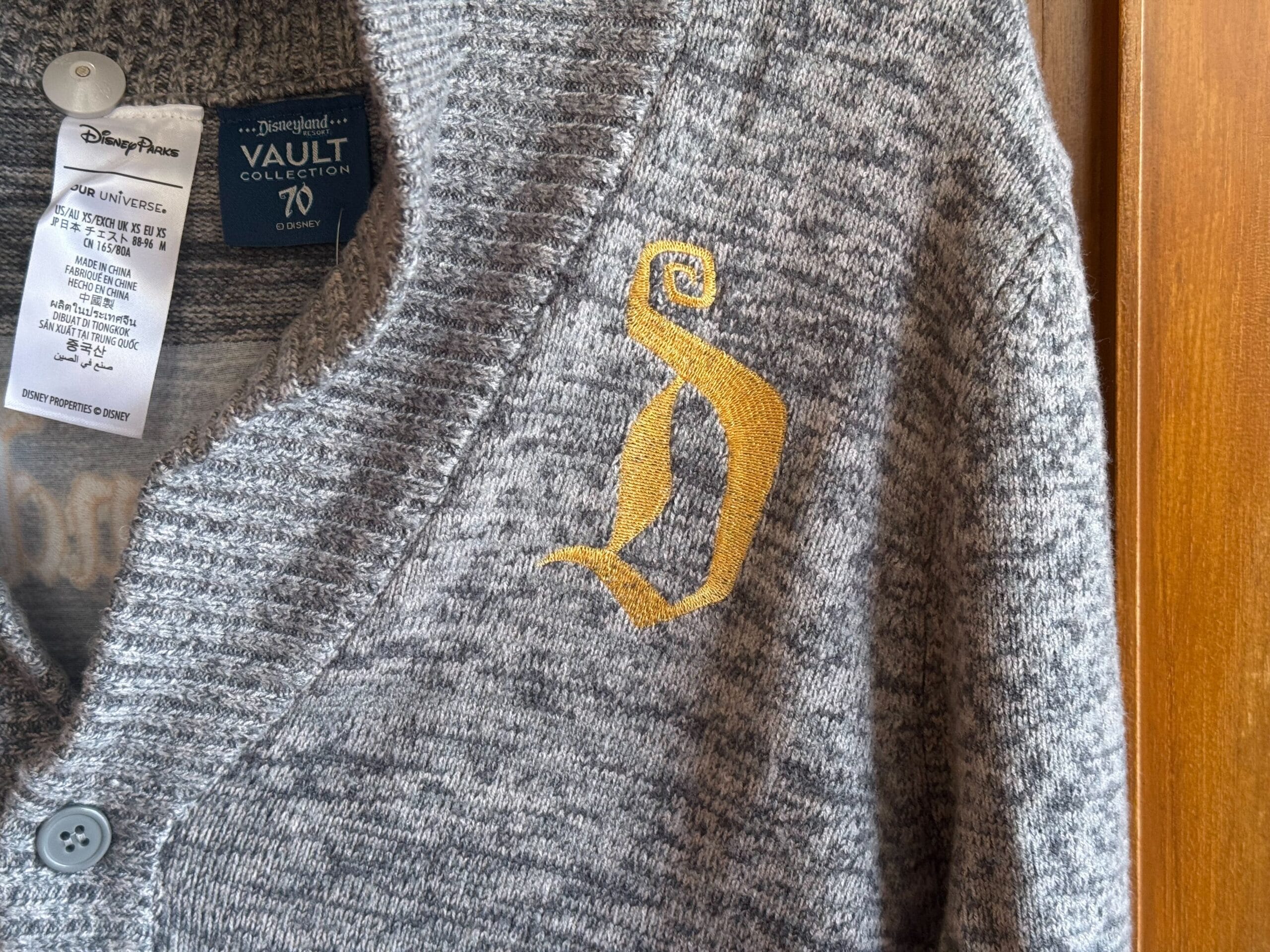 Grey cardigan with yellow Disney "D" embroidered, Vault Collection tag on collar—classic Disney Parks style, no castle in view.