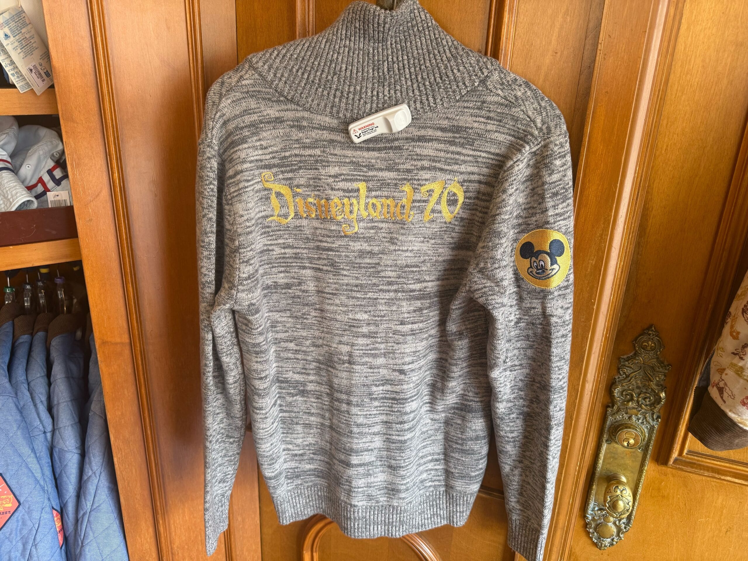 Gray sweater with "Disneyland 70" in gold on the back, Mickey Mouse patch on sleeve, hanging—classic park style, no castle view.