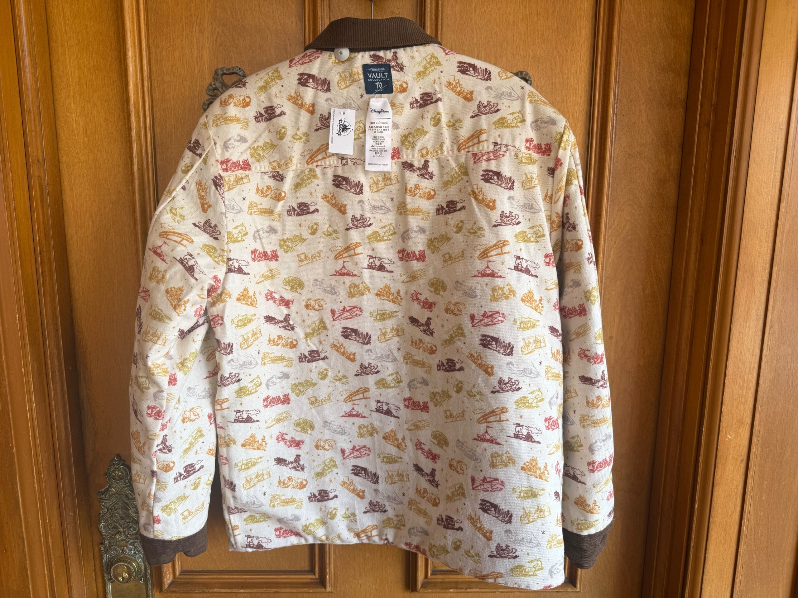A patterned jacket with colorful shoe prints, reminiscent of Disney parks flair, hangs on a wooden door, back and tags shown.