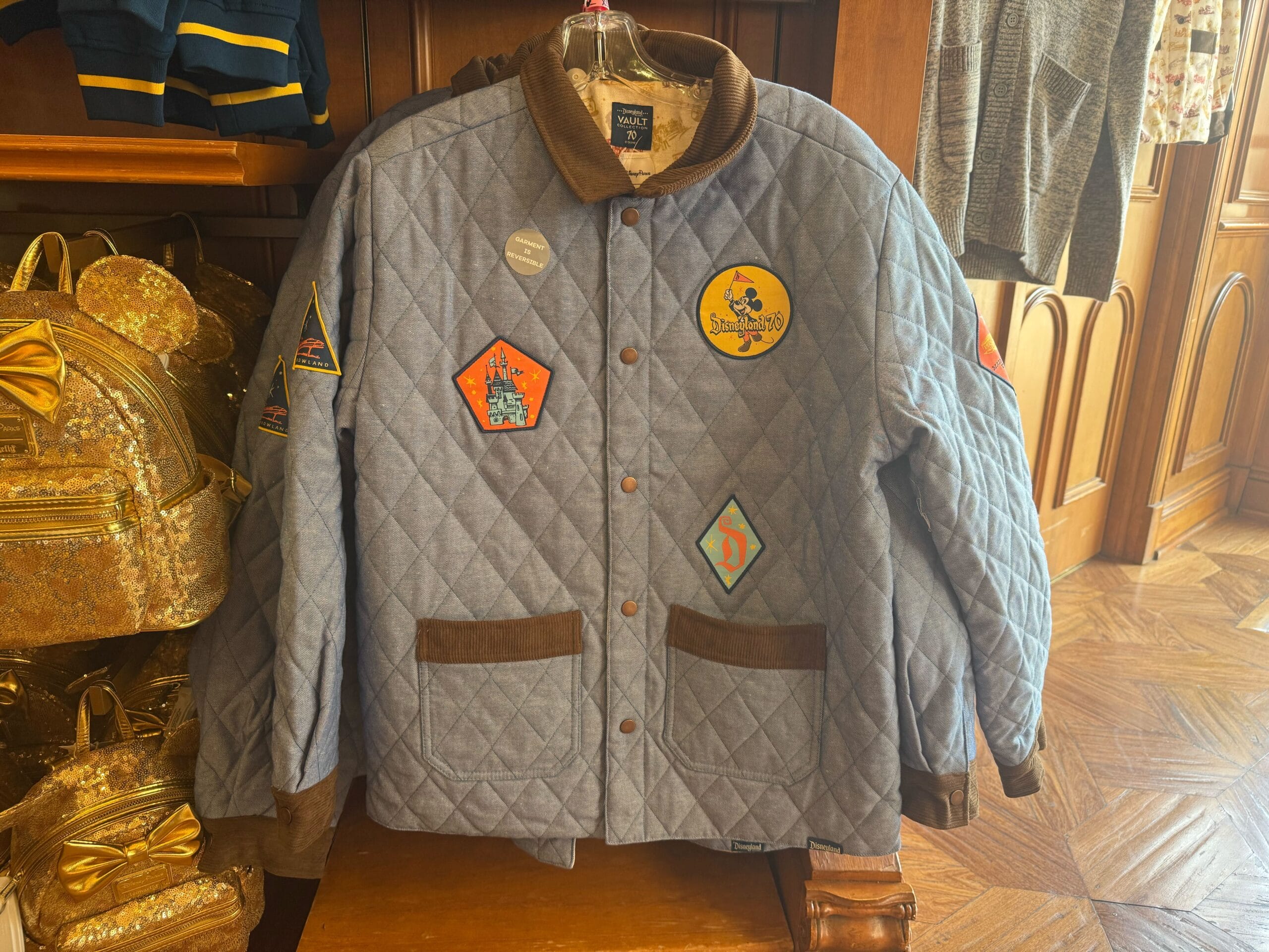 A quilted blue jacket with brown trim and colorful embroidered patches hangs on display in a Disney Park shop, seen near castle merch.