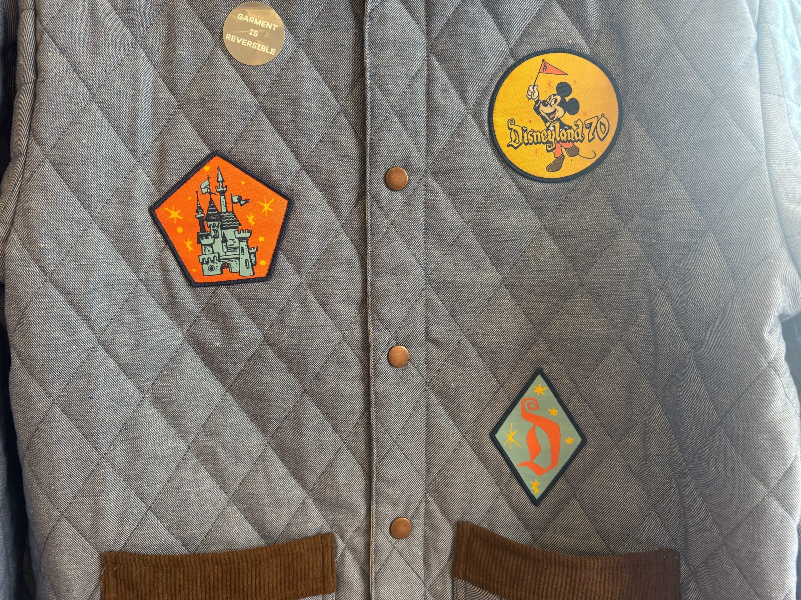 Quilted gray jacket featuring three vibrant Disneyland patches and a “GARMENT IS REVERSIBLE” button, reminiscent of park styles.
