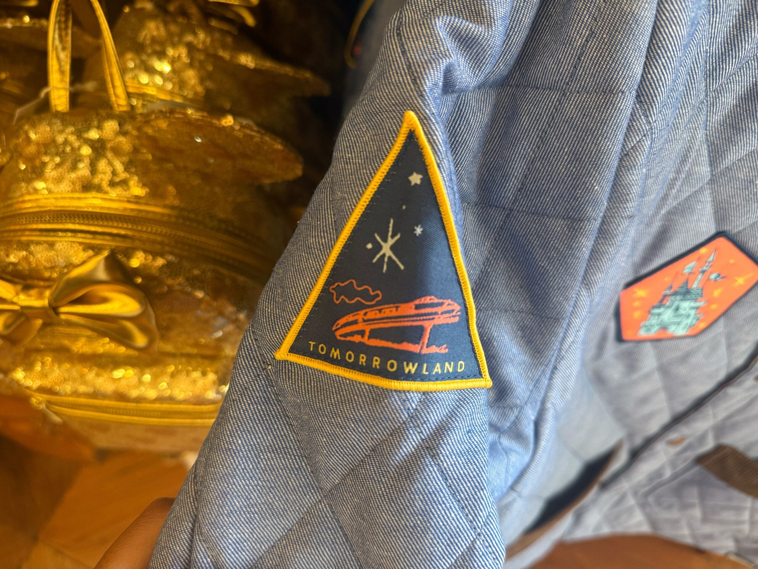 A blue Tomorrowland jacket with a spaceship-and-stars patch, reminiscent of Disney parks, beside a gold backpack.