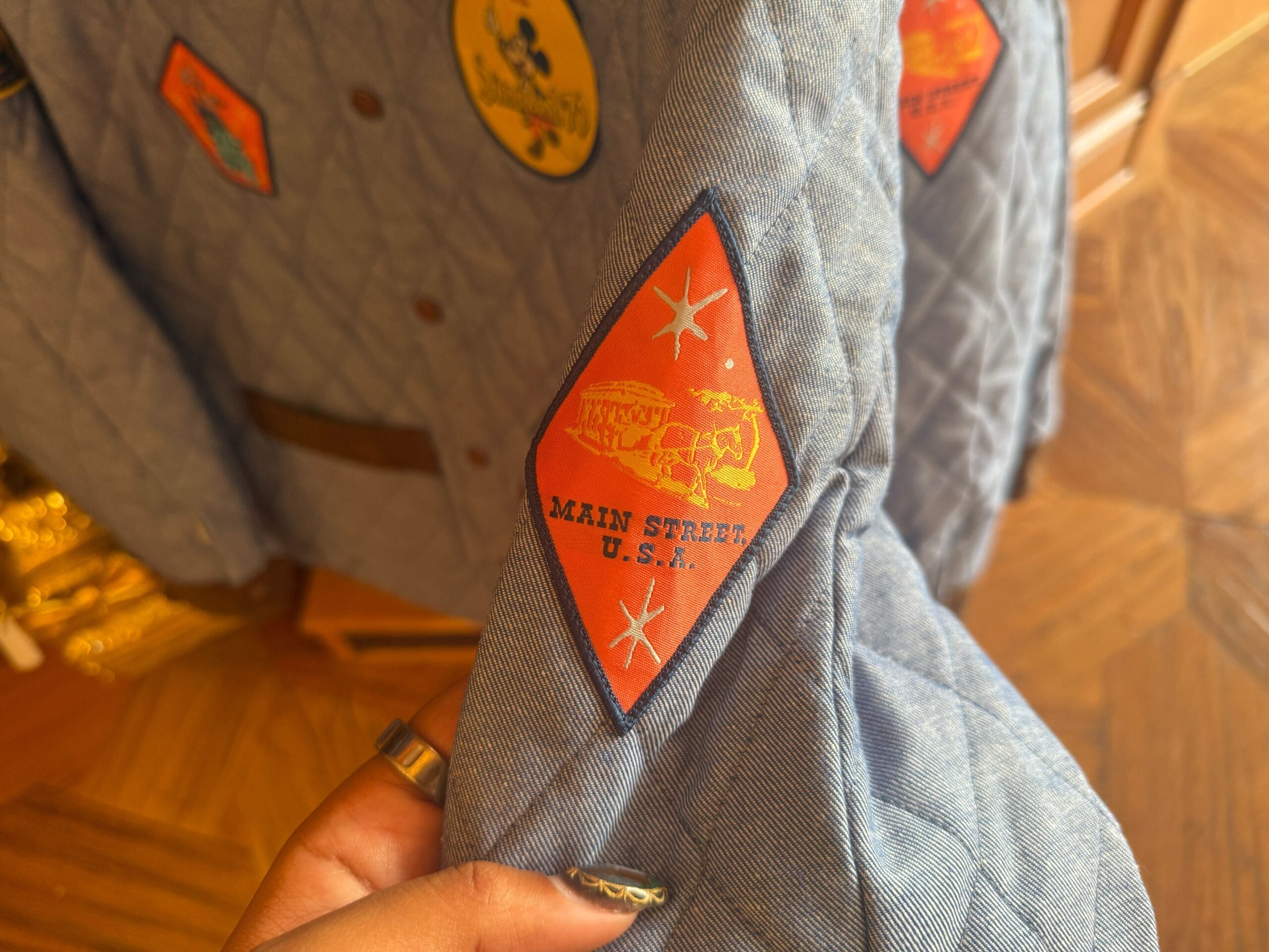 A hand displays a blue jacket sleeve with a red “Main Street U.S.A.” patch and map motif, inspired by the classic Disney park entrance.