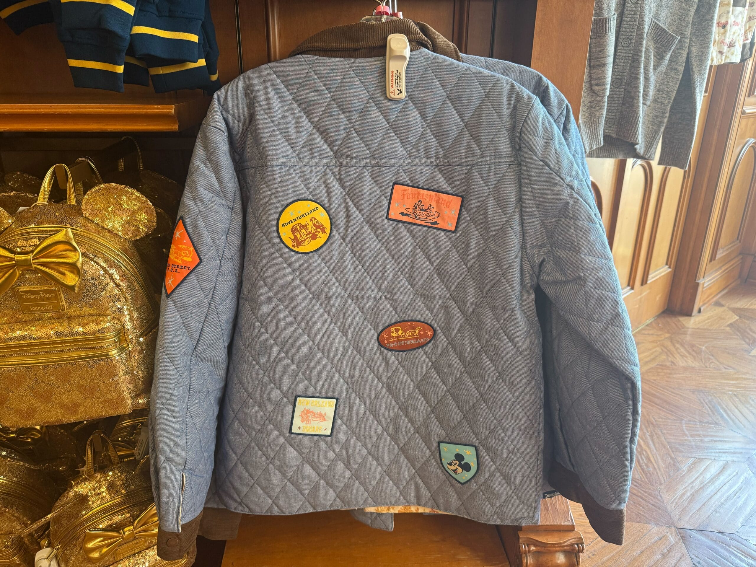 A blue quilted jacket decorated with vibrant patches hangs near a gold glittery backpack, like finds from a Disney Park shop.