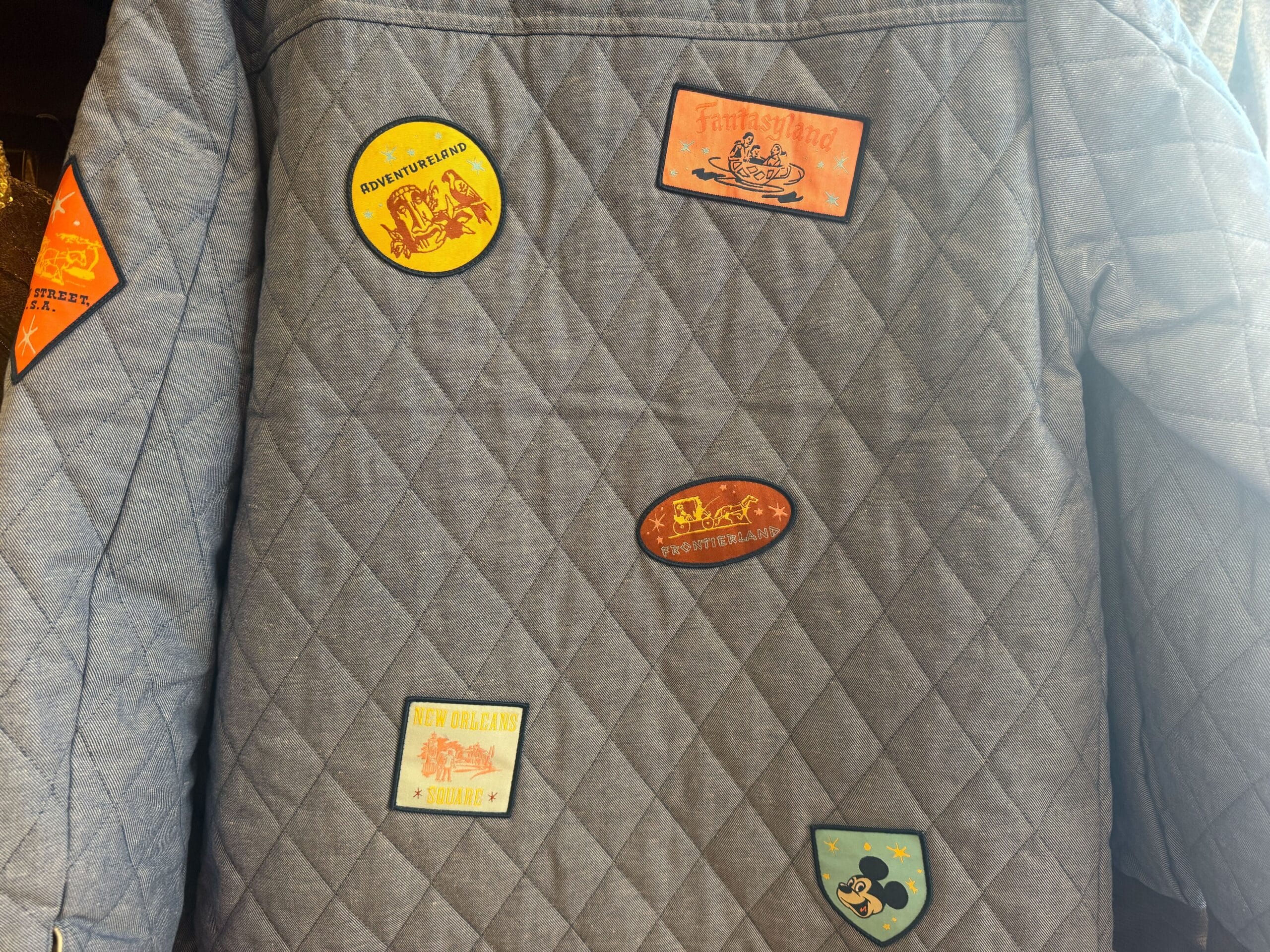 A quilted gray jacket with six colorful embroidered patches, each showcasing themes inspired by Disney Parks attractions and icons.