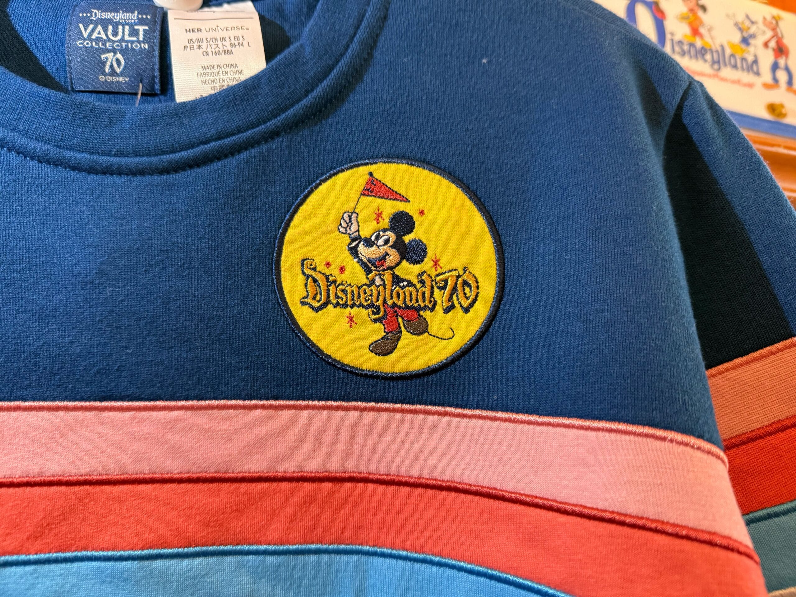Blue sweatshirt with Disneyland 70 patch showing Mickey Mouse holding a flag, above pink and blue stripes; no castle present.