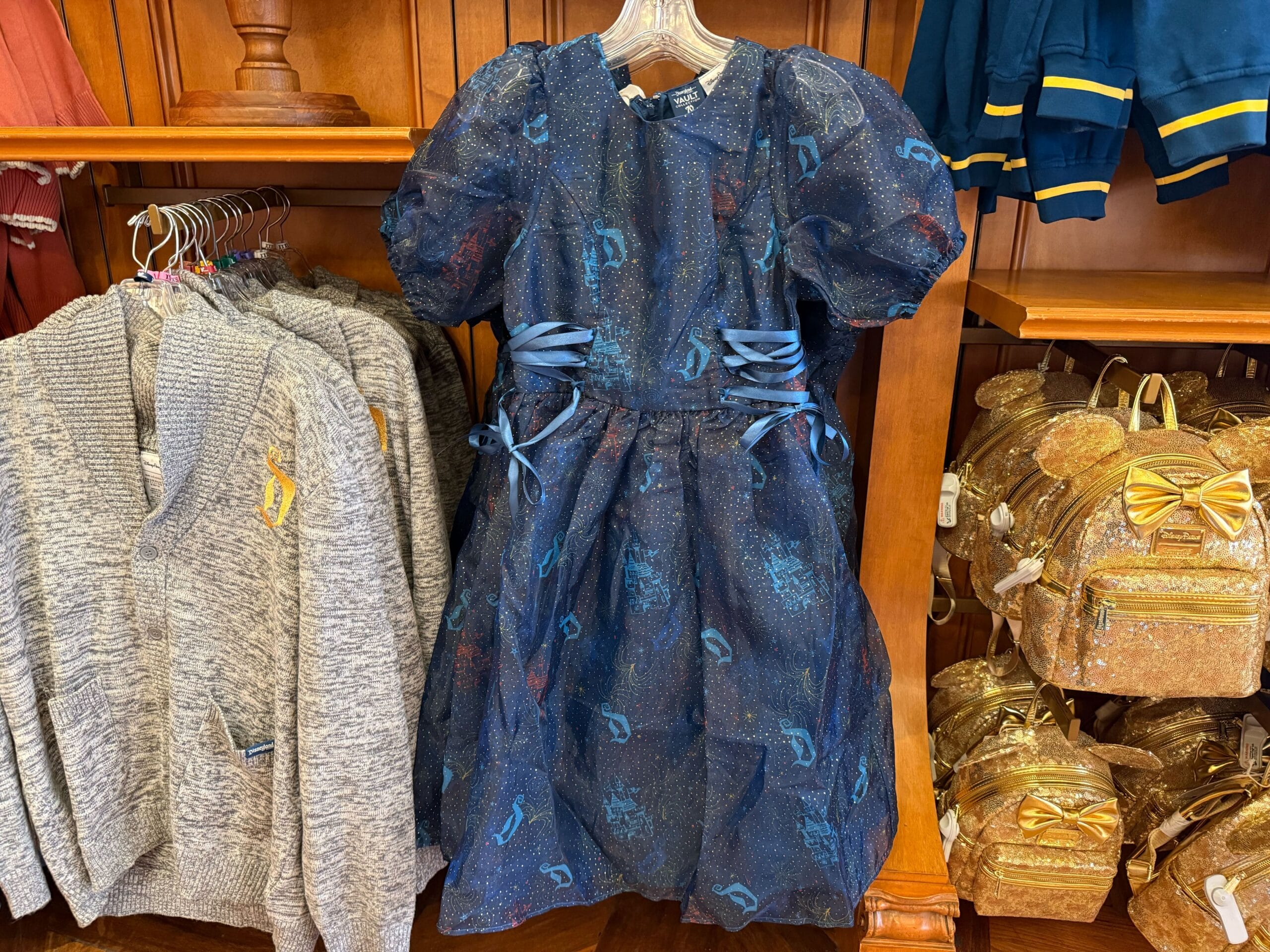 Sparkly navy blue girl's dress with puff sleeves and blue ribbons, similar to Disney princess style, next to sweaters and gold backpacks.