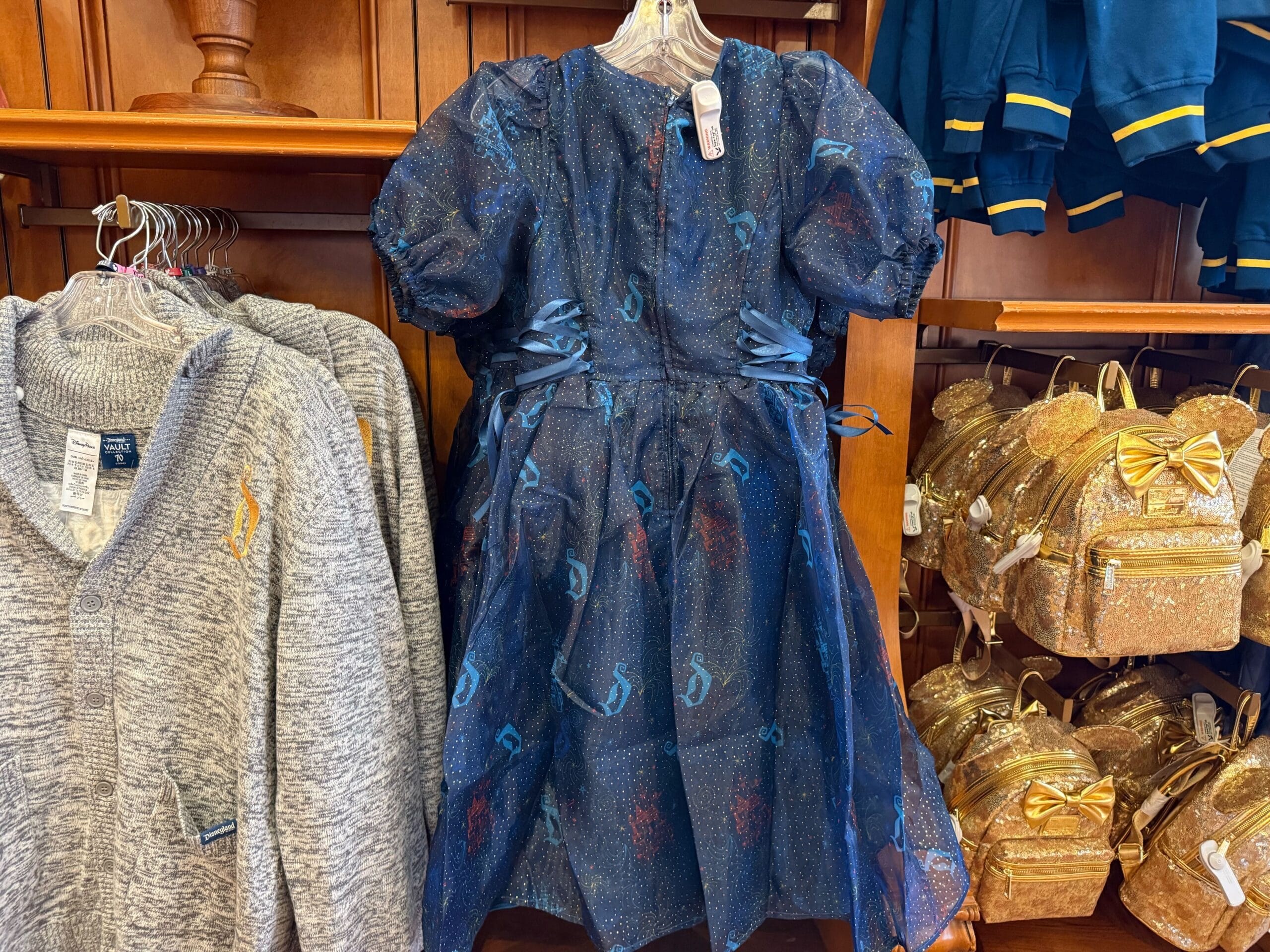 A blue sparkly dress with Hogwarts house crests hangs by sweaters and golden backpacks, resembling Disney park merchandise displays.
