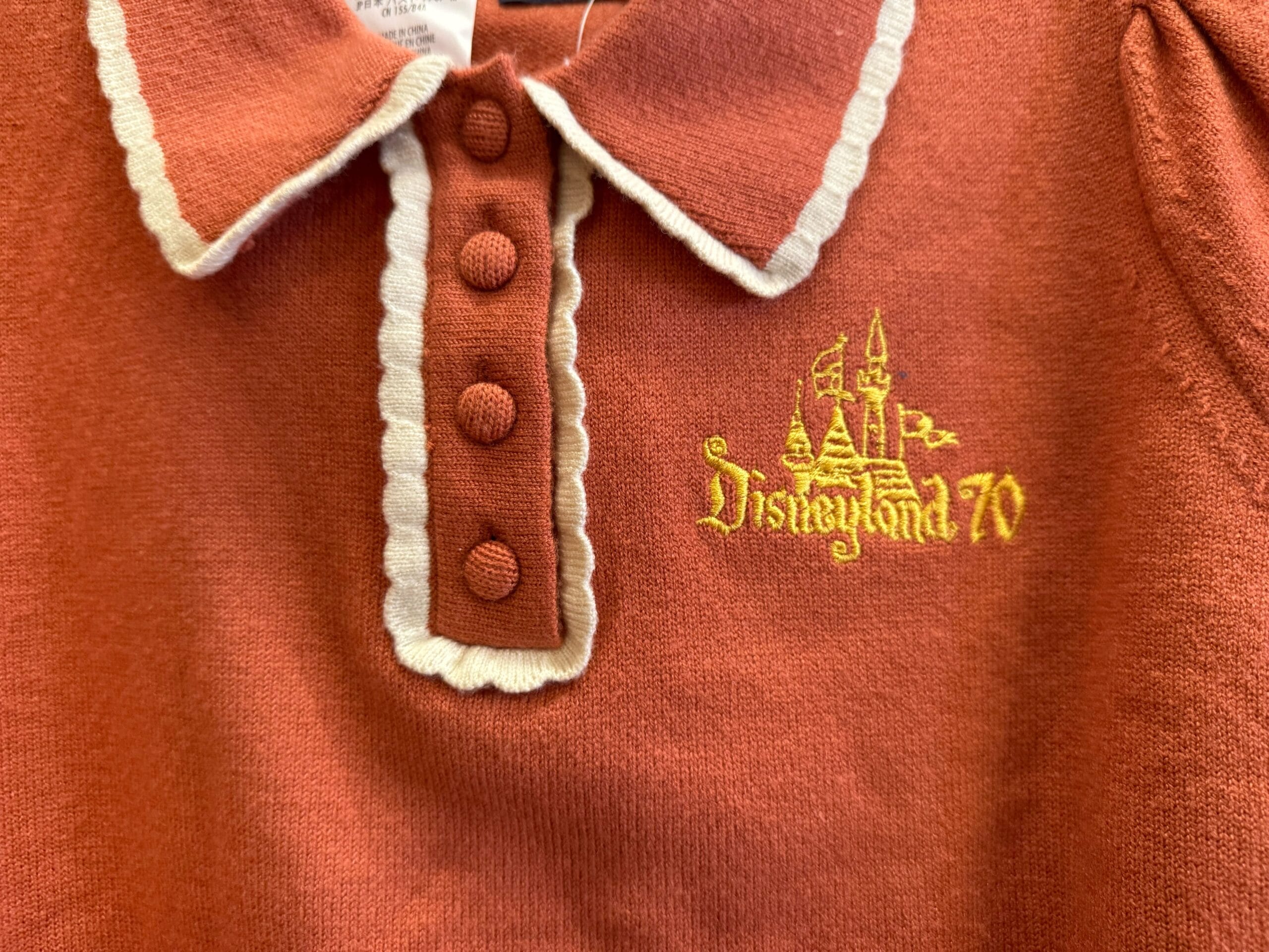 Rust shirt with white trim and buttons, plus a yellow "Disneyland 70" logo showing a castle reminiscent of the Anaheim park’s design.