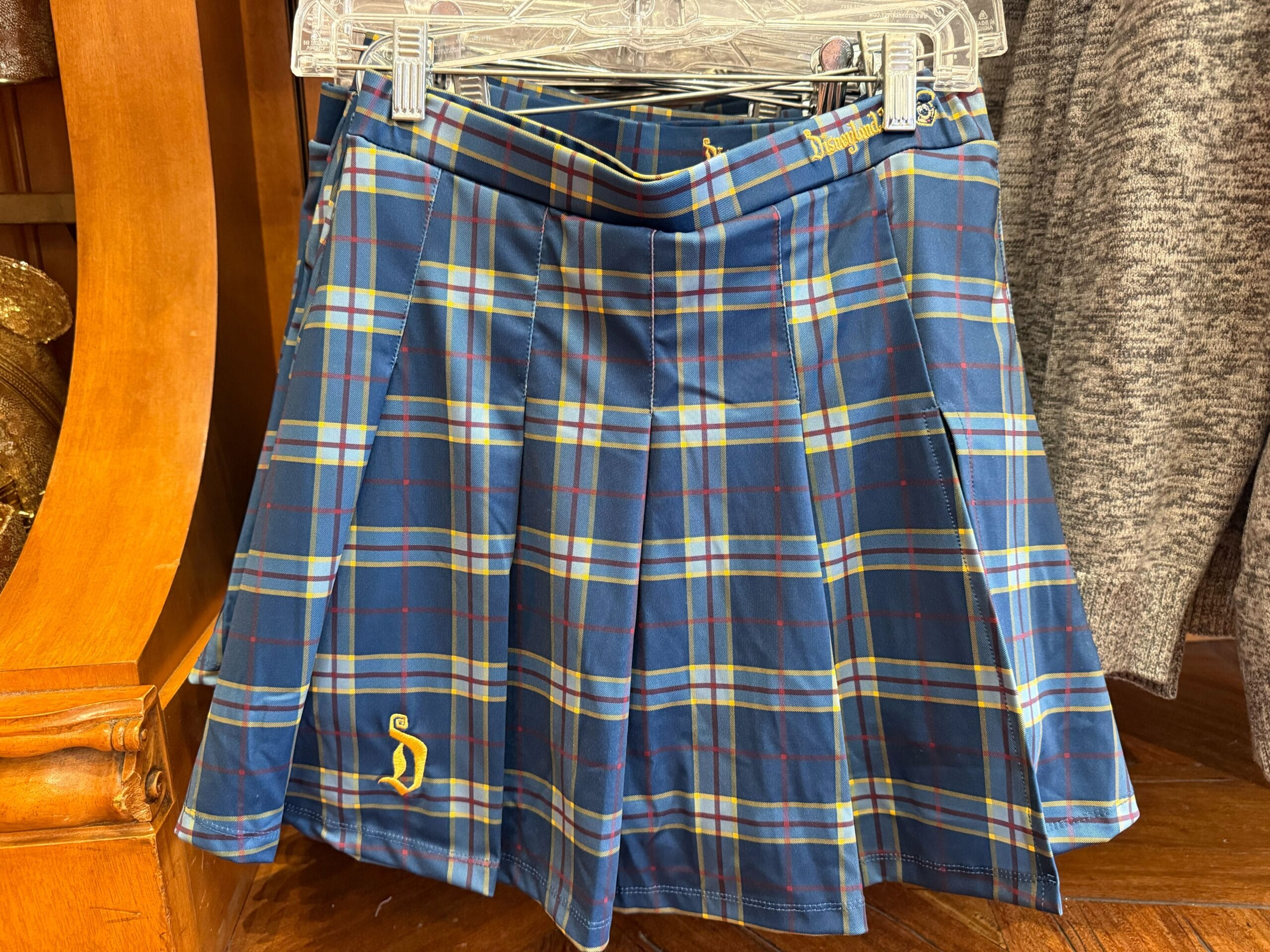 Blue and green plaid pleated skirt on a hanger, featuring a yellow "S" near the hem—evokes classic Disney School Spirit styles.