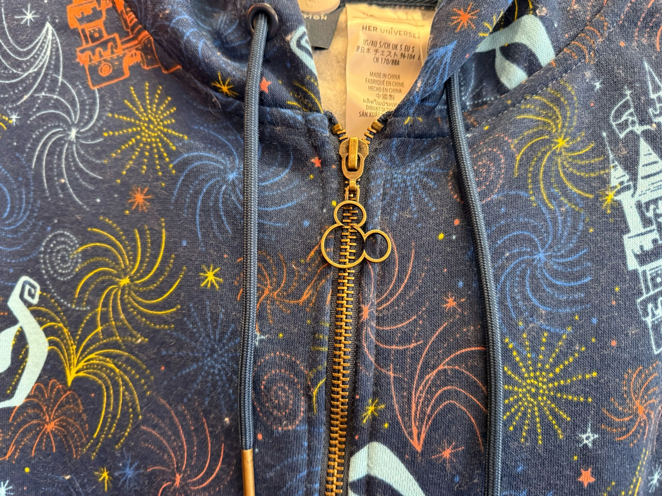 Close-up of a hoodie zipper with a Mickey Mouse pull, colorful fireworks, and a castle reminiscent of Disneyland's Sleeping Beauty Castle.