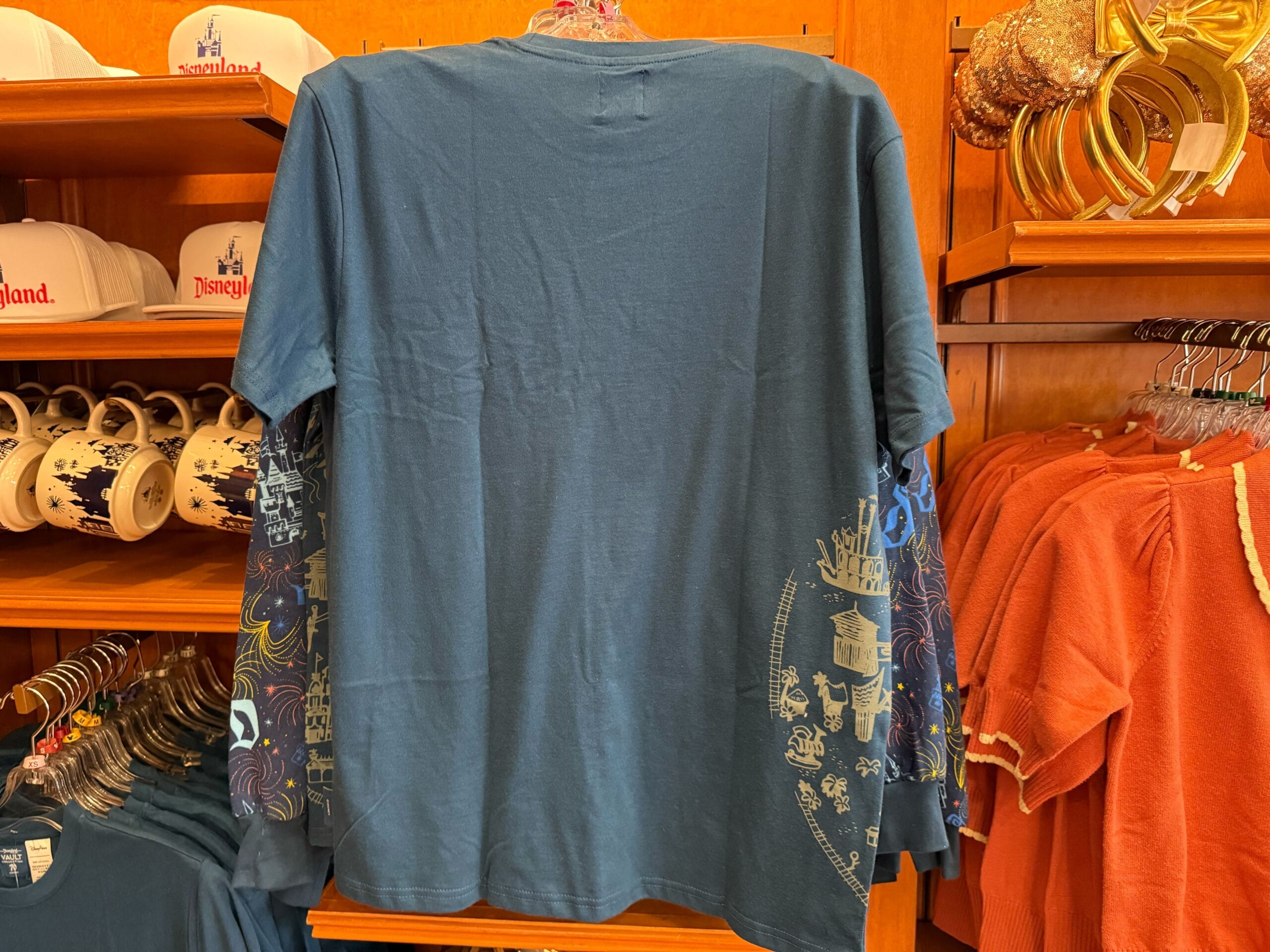 Blue long-sleeve T-shirt featuring gold and blue ride icons from Disneyland on sleeves and lower back, displayed in a Disney park shop.