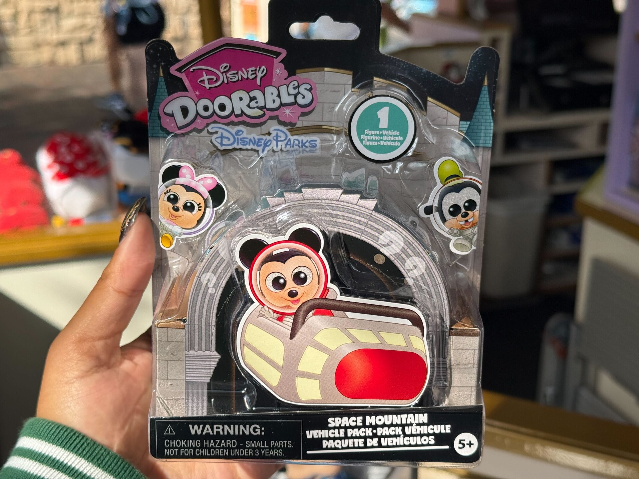 Hand holding a Disney Doorables Space Mountain vehicle toy pack with Mickey, park background resembles Disneyland, not Cinderella Castle.