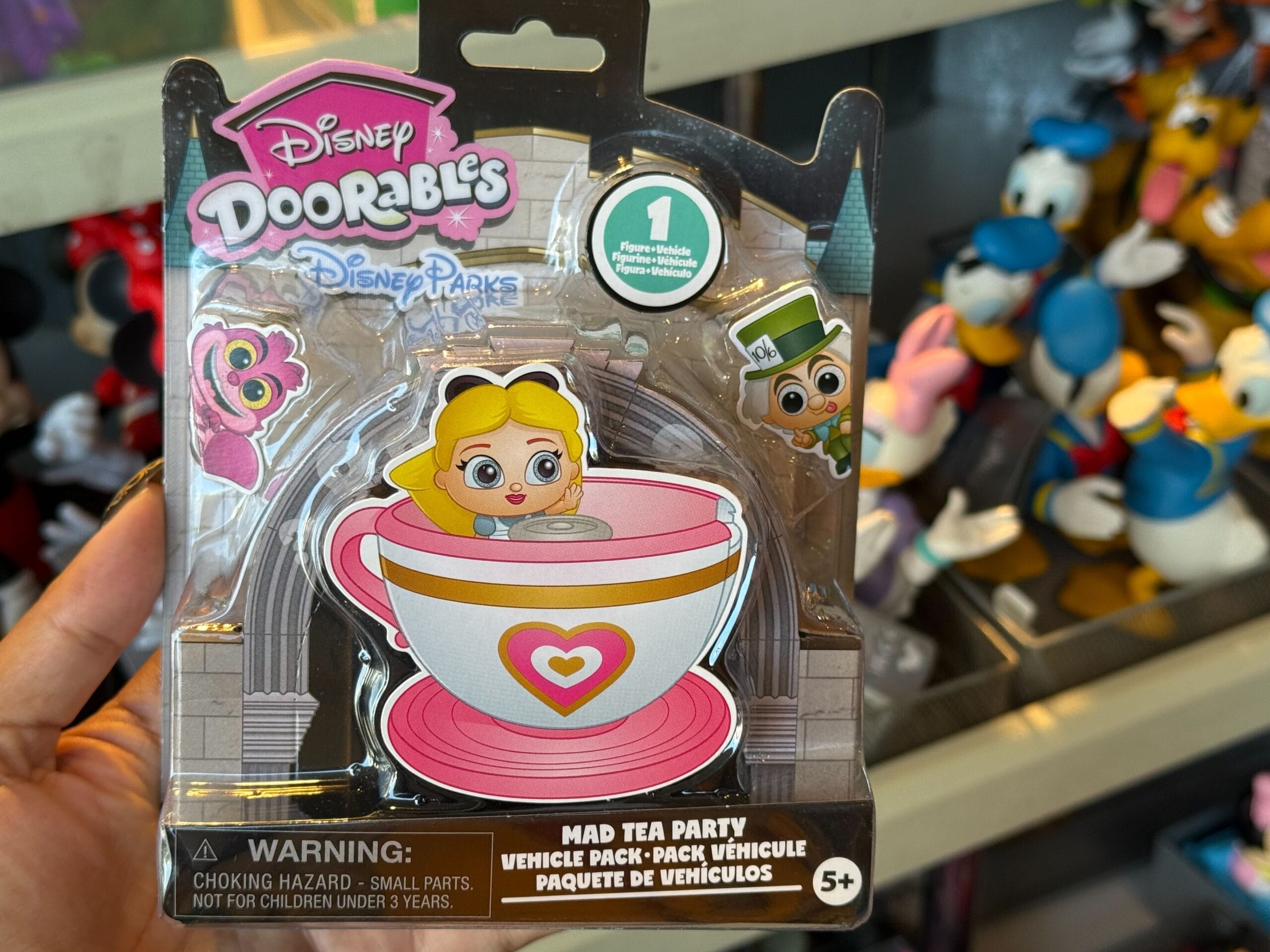 A hand holds a Disney Doorables "Mad Tea Party" toy pack with a figure in a pink teacup, inspired by the iconic park attraction.