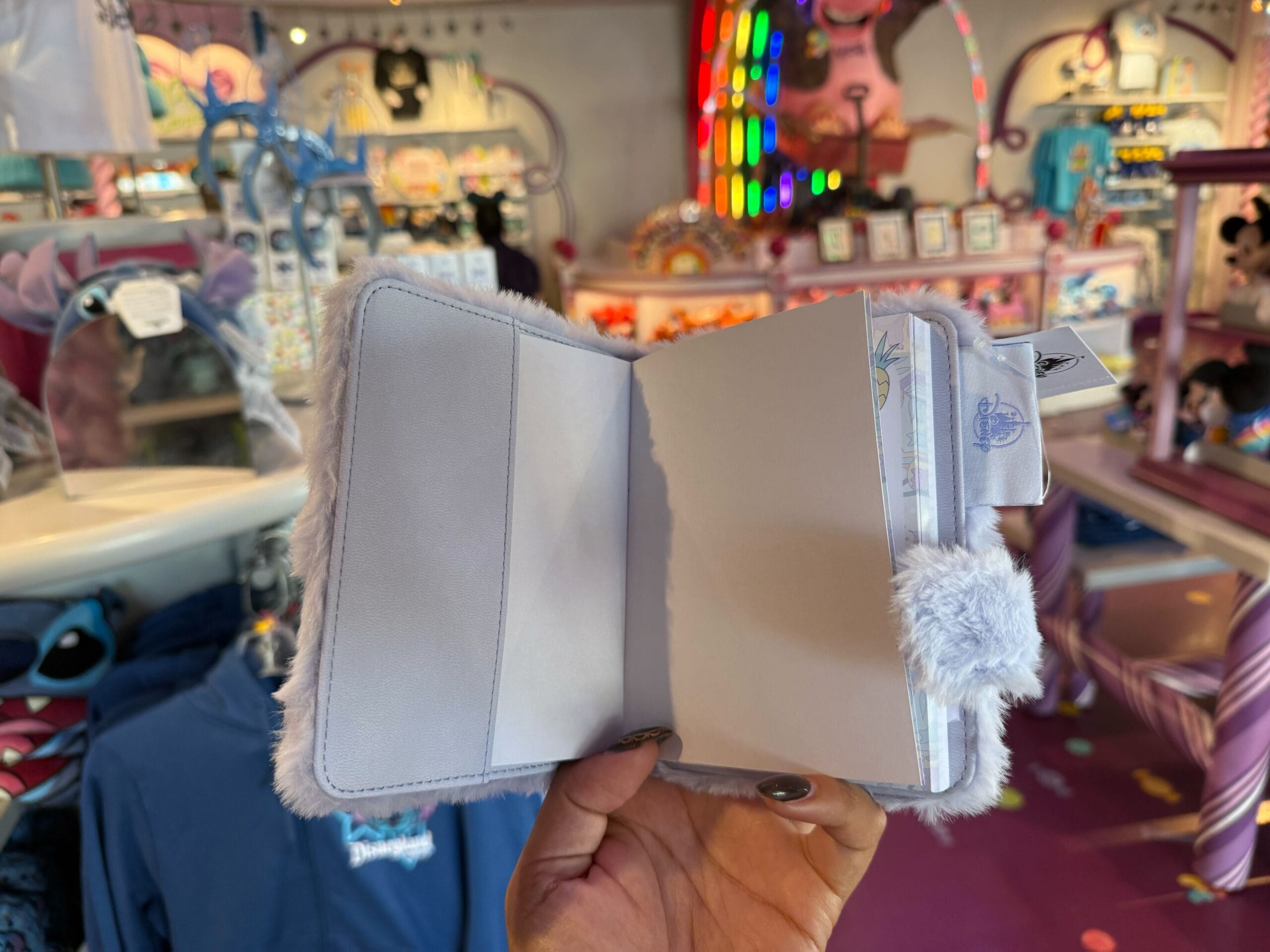 A hand displays a Fluffy Stitch Journal with blank pages inside a vibrant Disney park shop brimming with Stitch merchandise.
