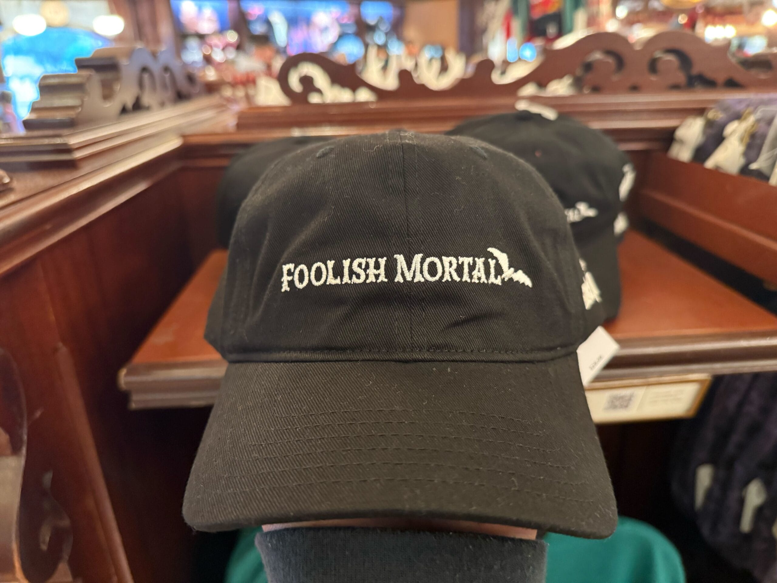 A black baseball cap with "Foolish Mortal" in white on the front, reminiscent of Disney's Haunted Mansion, sits on a wooden shelf.