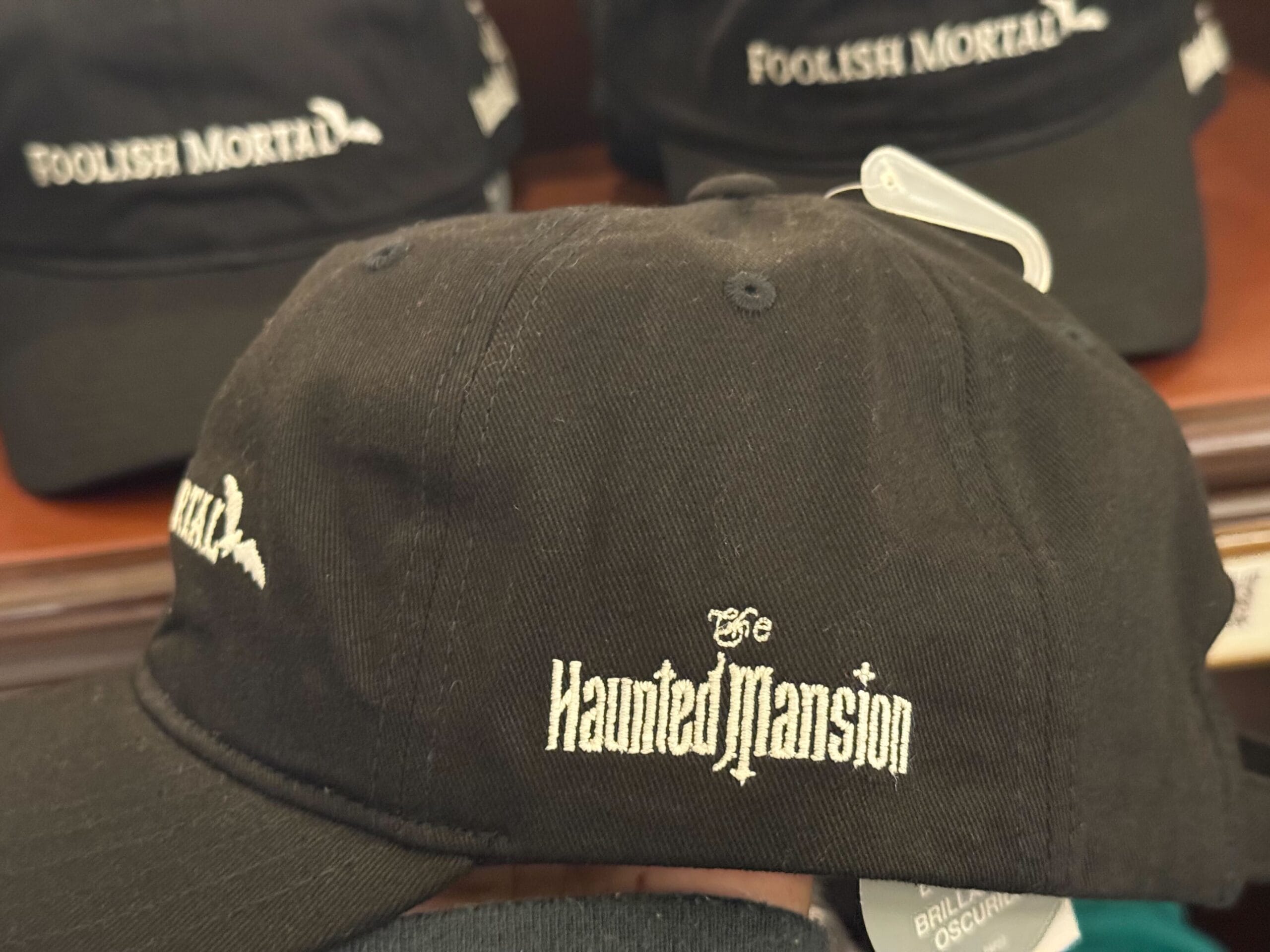 Black cap with "The Haunted Mansion" embroidery, surrounded by "Foolish Mortal" caps—a staple from Disney Parks merchandise.