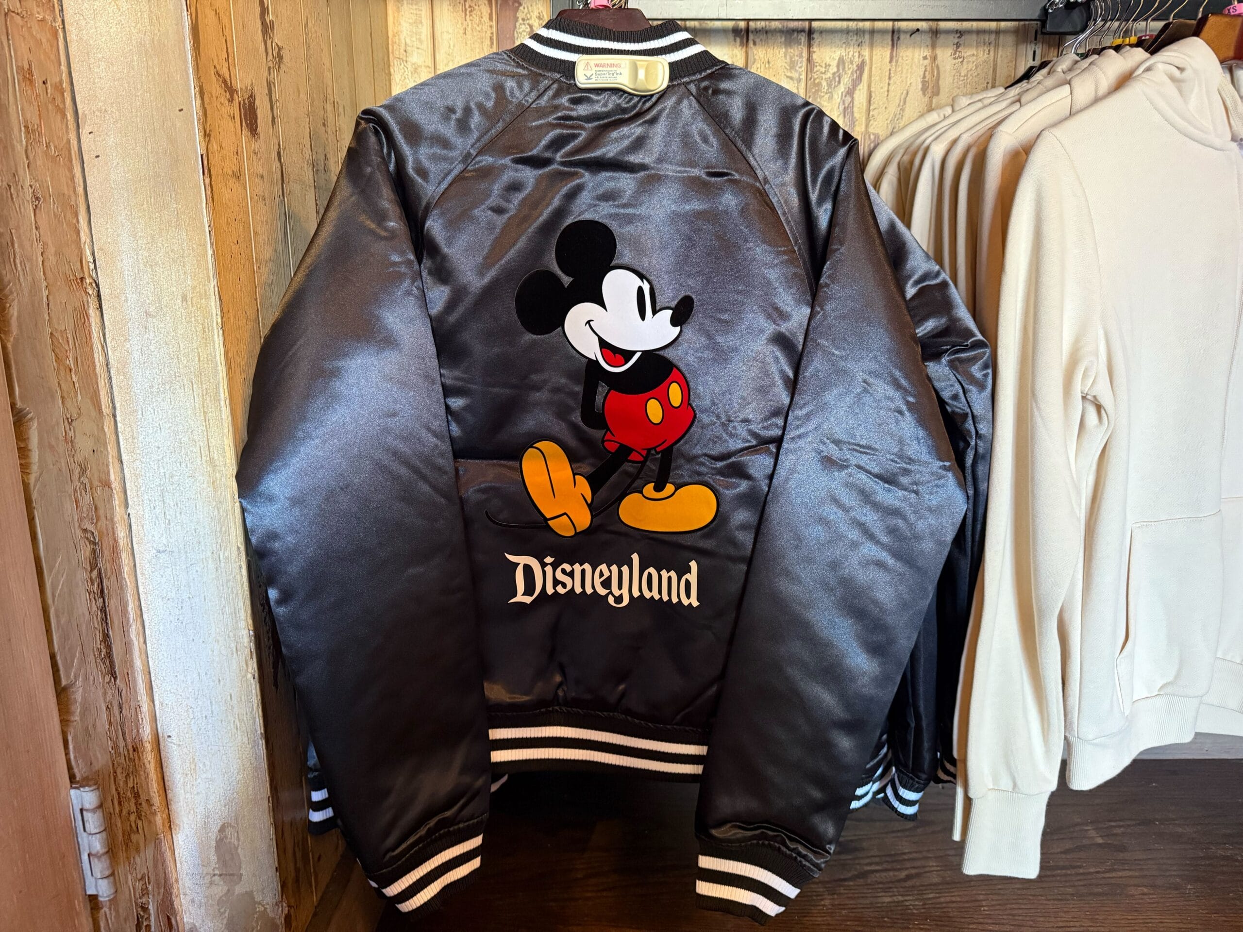 Black satin Disneyland jacket featuring Mickey Mouse on the back, hanging beside beige hoodies, likely in a park gift shop.