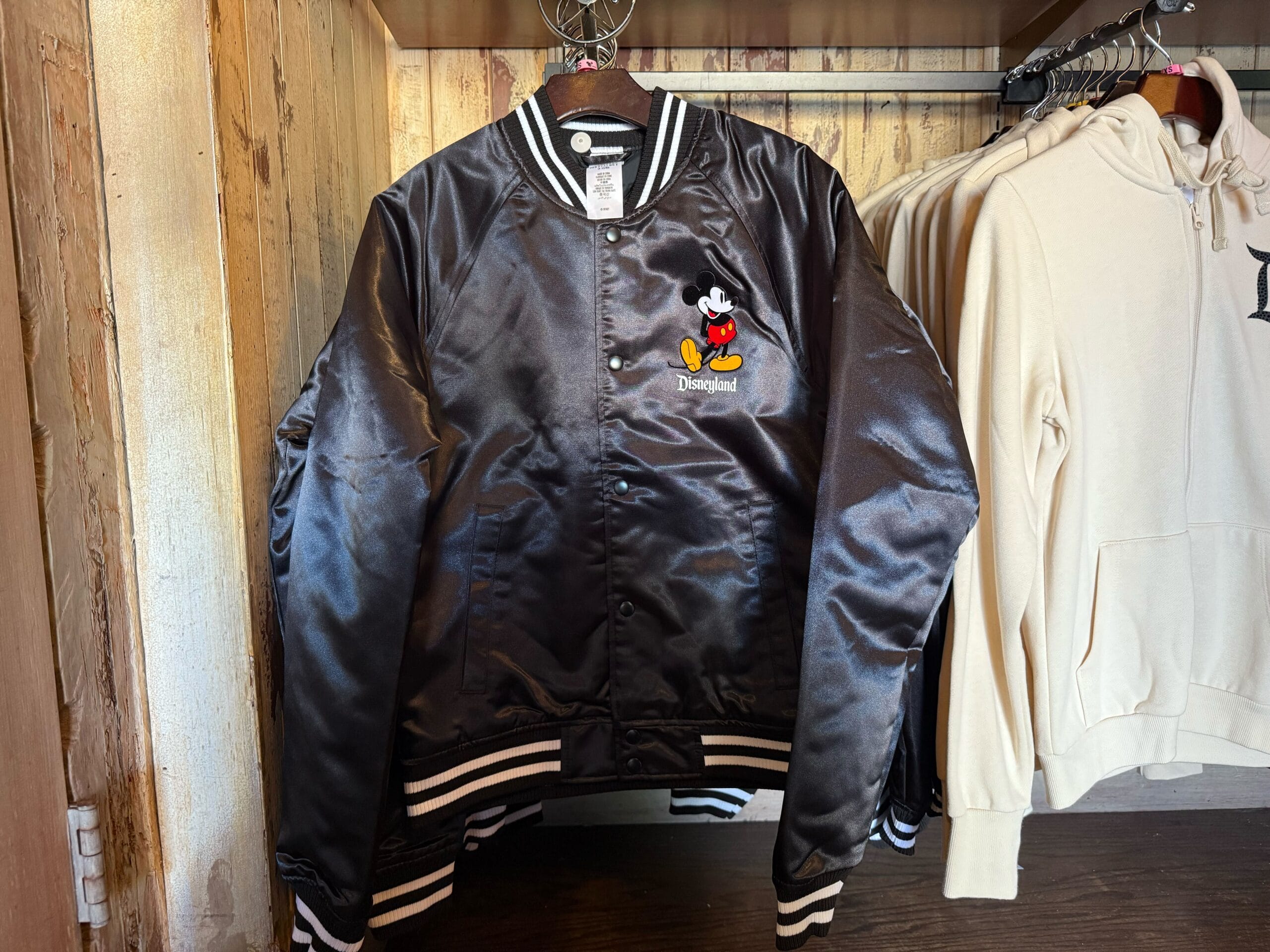 A black satin Disneyland jacket with a Mickey Mouse patch hangs beside cream hoodies, reminiscent of park shops near castle entrances.