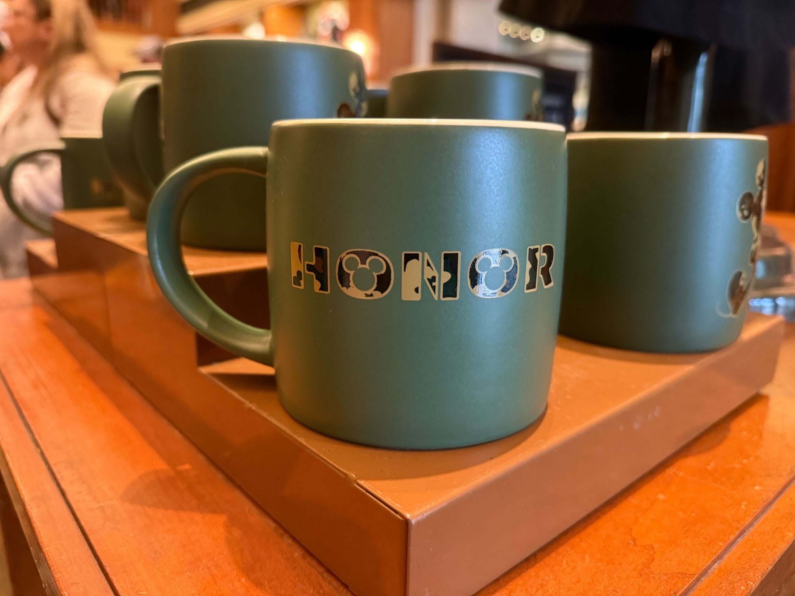 A green mug featuring “HONOR” in black and gold Disney camouflage print letters with a Mickey head in the O.