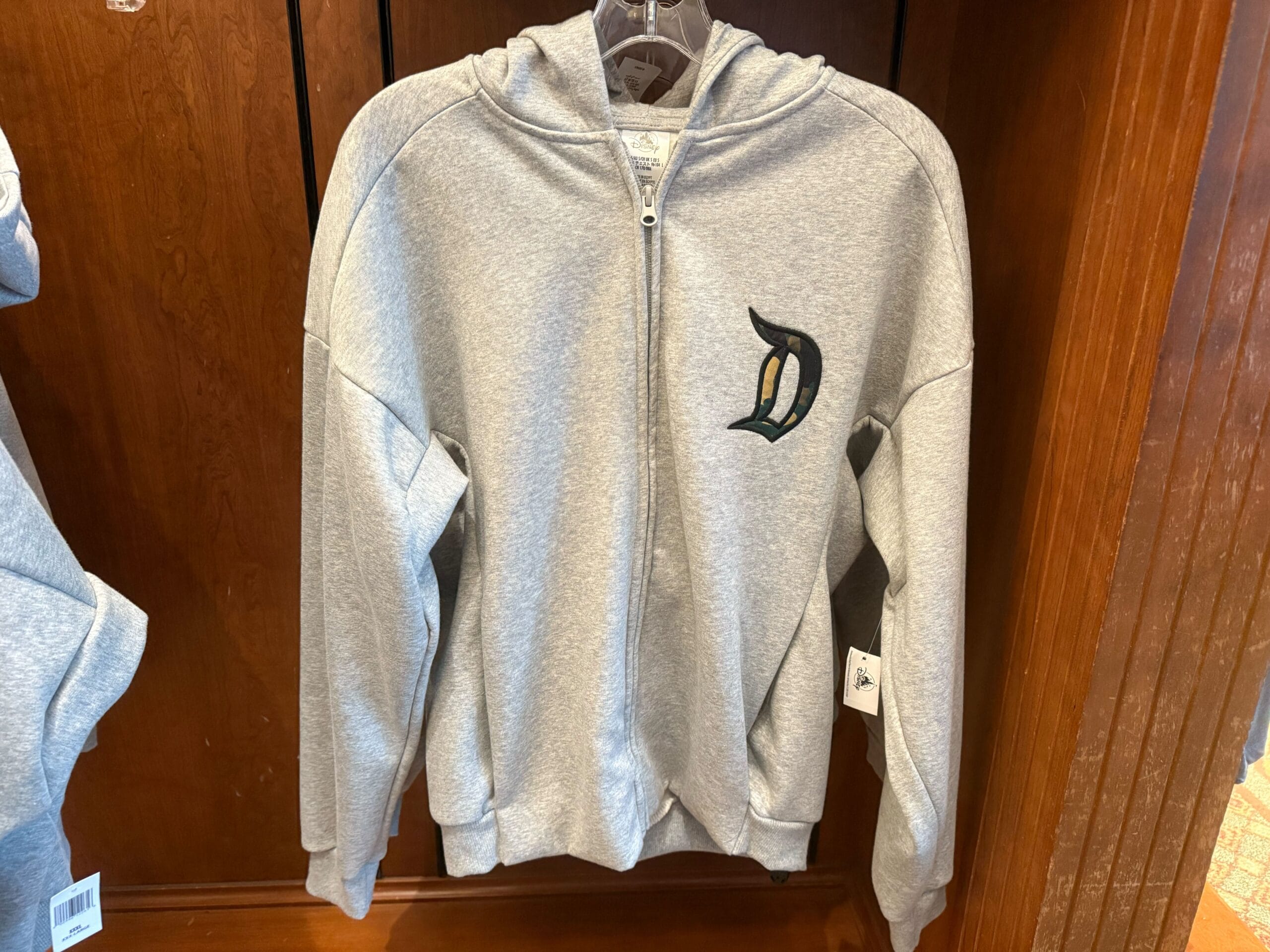 Gray zip-up hoodie with a Disney-style "D" on the chest in camouflage print outlined in black stitching, displayed on a wooden rack.