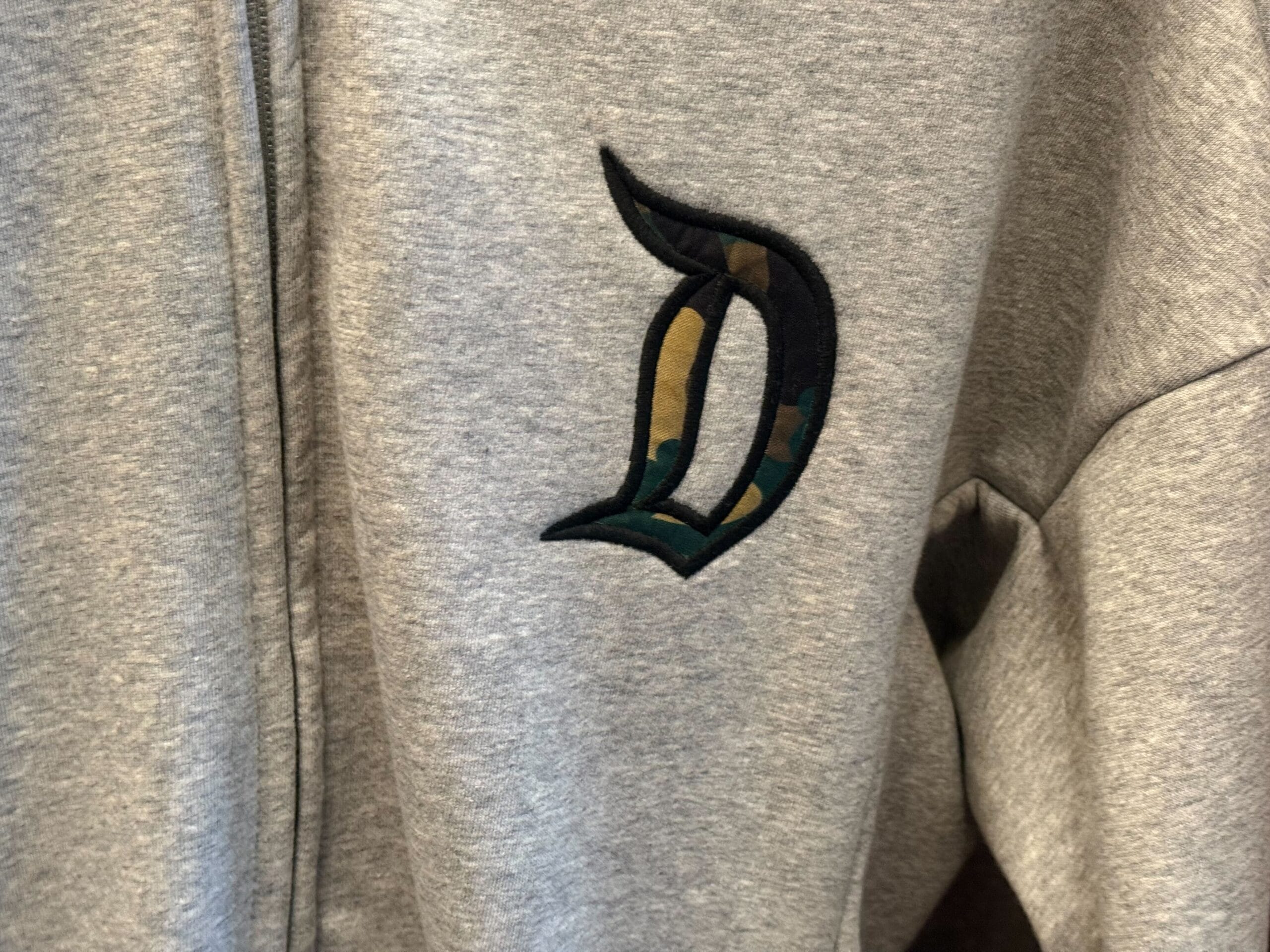 Close up of a gray zip-up sweatshirt featuring a large green camouflage Disneyland "D" appliqued on the front left chest.