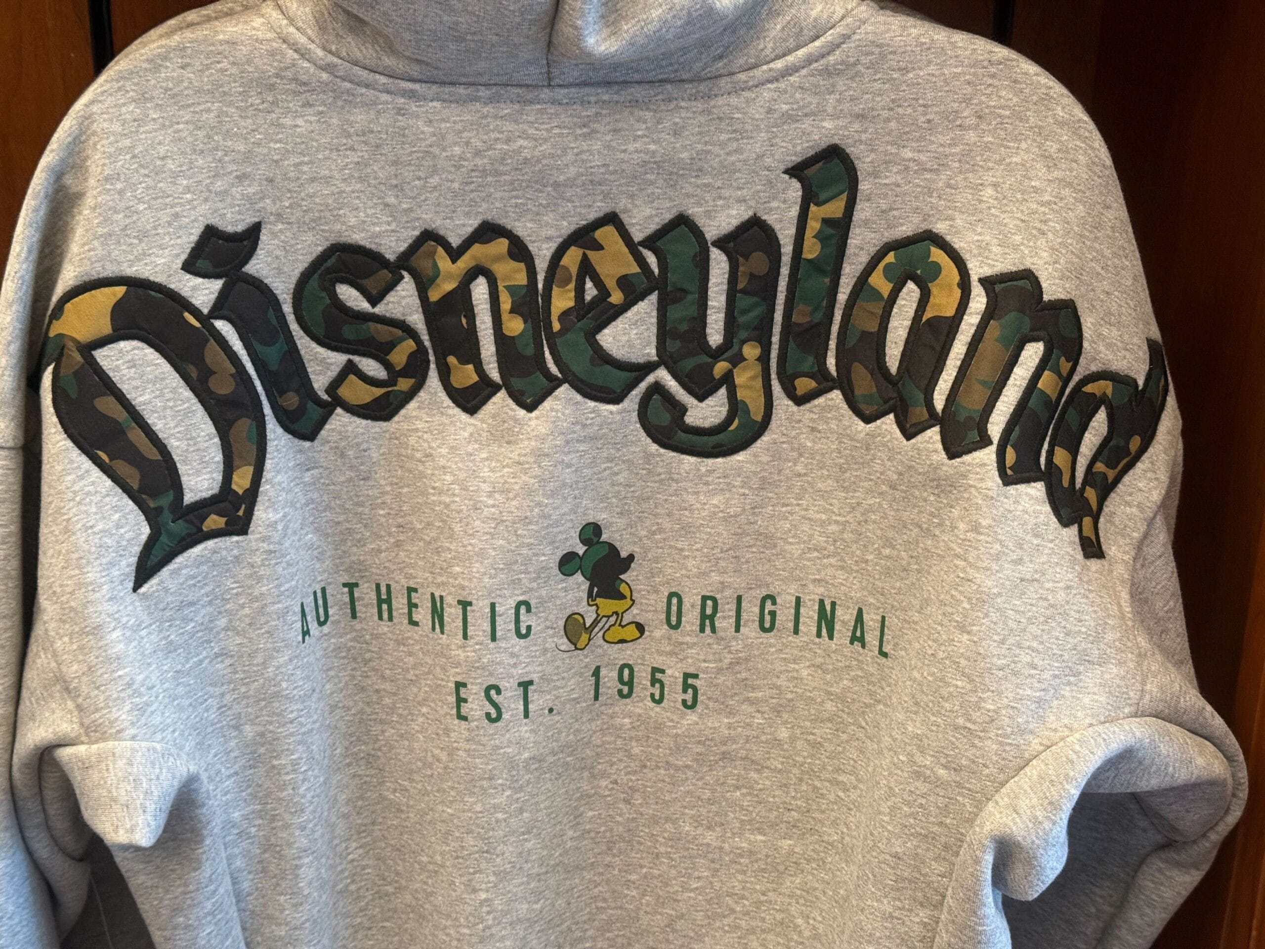 Back of gray Disneyland hoodie featuring bold Mickey camouflage print “Disneyland” lettering, plus “Authentic Original Est. 1955” on the back and a camouflage Mickey silhouette.