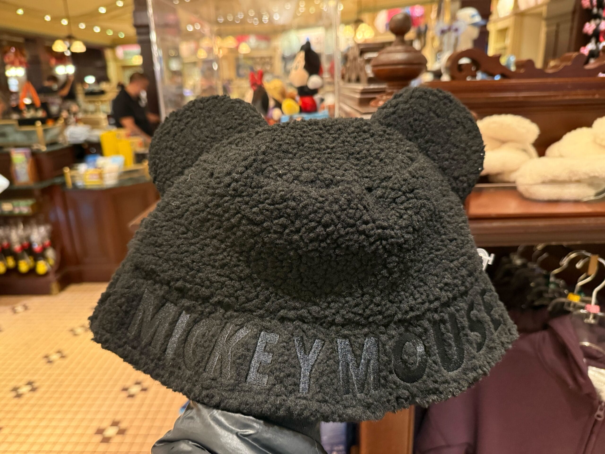 A soft black Sherpa cap with Mickey ears and "Mickey Mouse" on the front, seen in a Disneyland shop near Sleeping Beauty Castle.