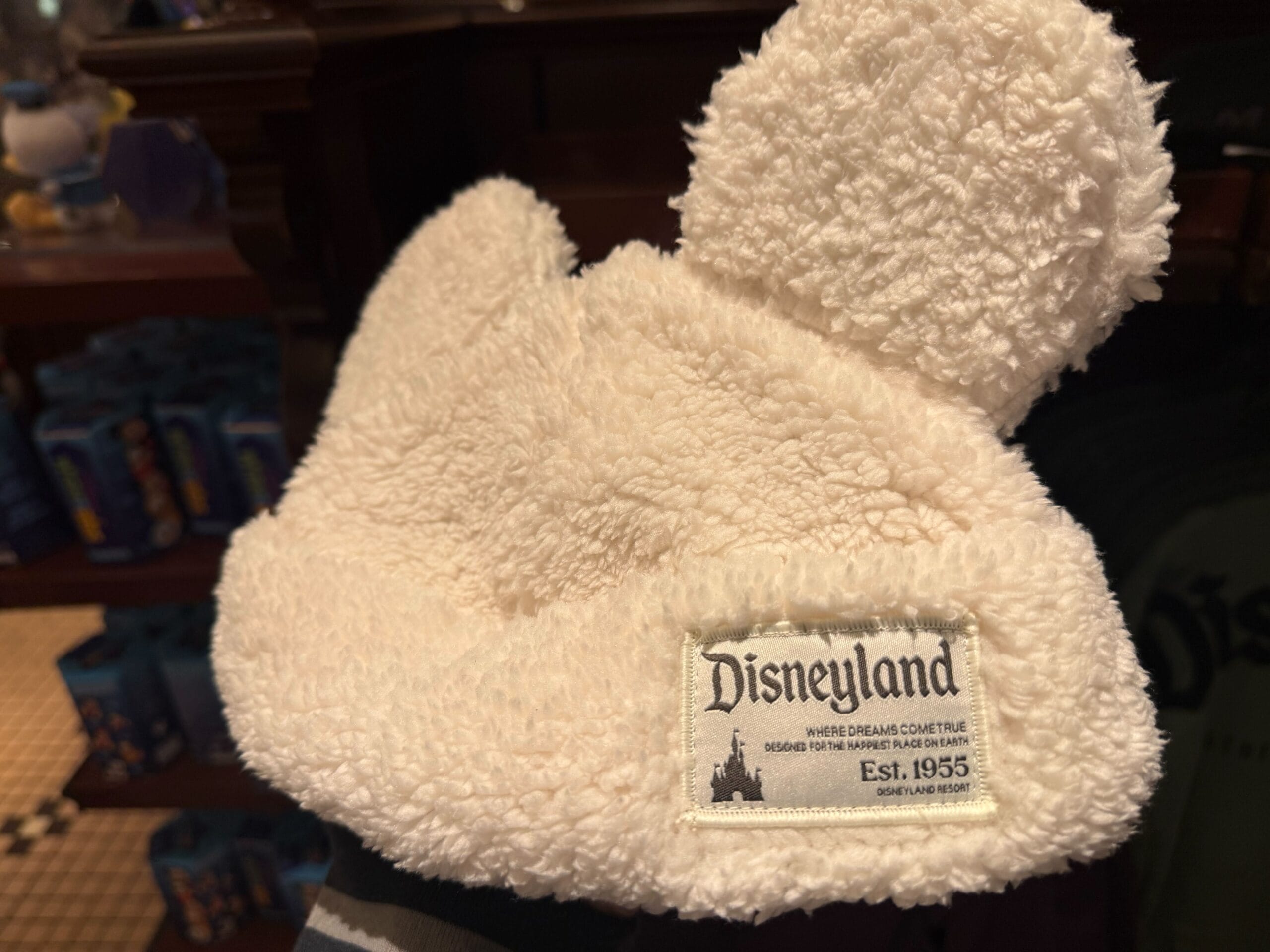 A fluffy white Sherpa cap with classic Mickey Mouse ears and a "Disneyland Est. 1955" tag, nodding to the original Anaheim park.