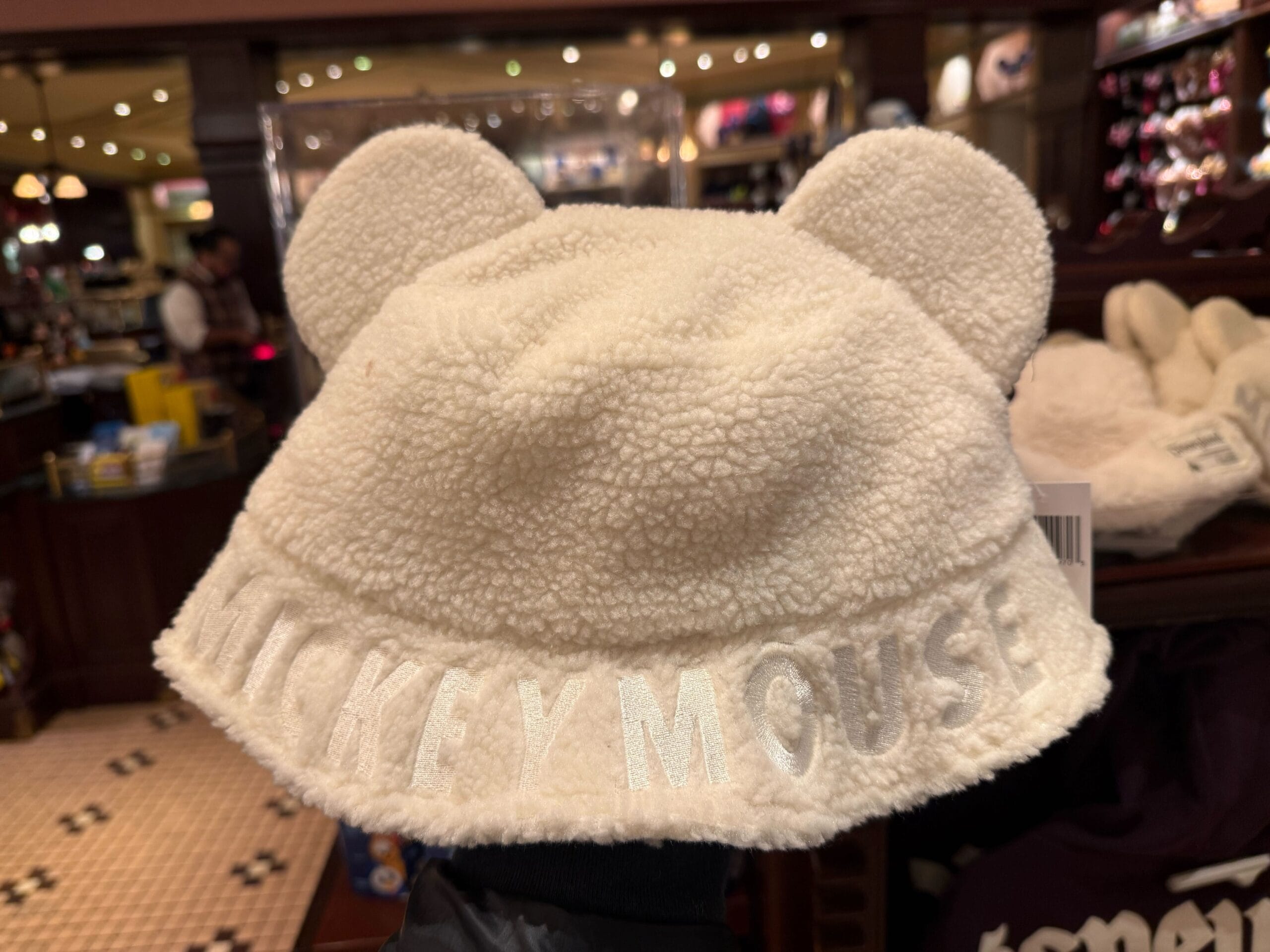 A fuzzy white Sherpa bucket hat with bear ears and "Mickey Mouse" stitched on front, spotted at a Disney Park store display.