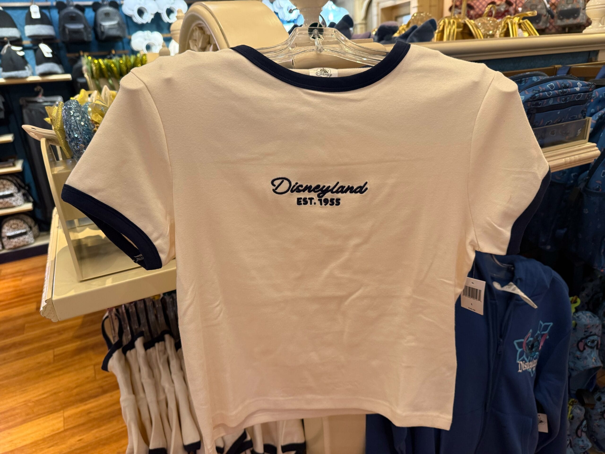 White Disneyland tee with navy trim, "Disneyland EST. 1955" chest embroidery, seen in a park shop—classic Anaheim style, not WDW.