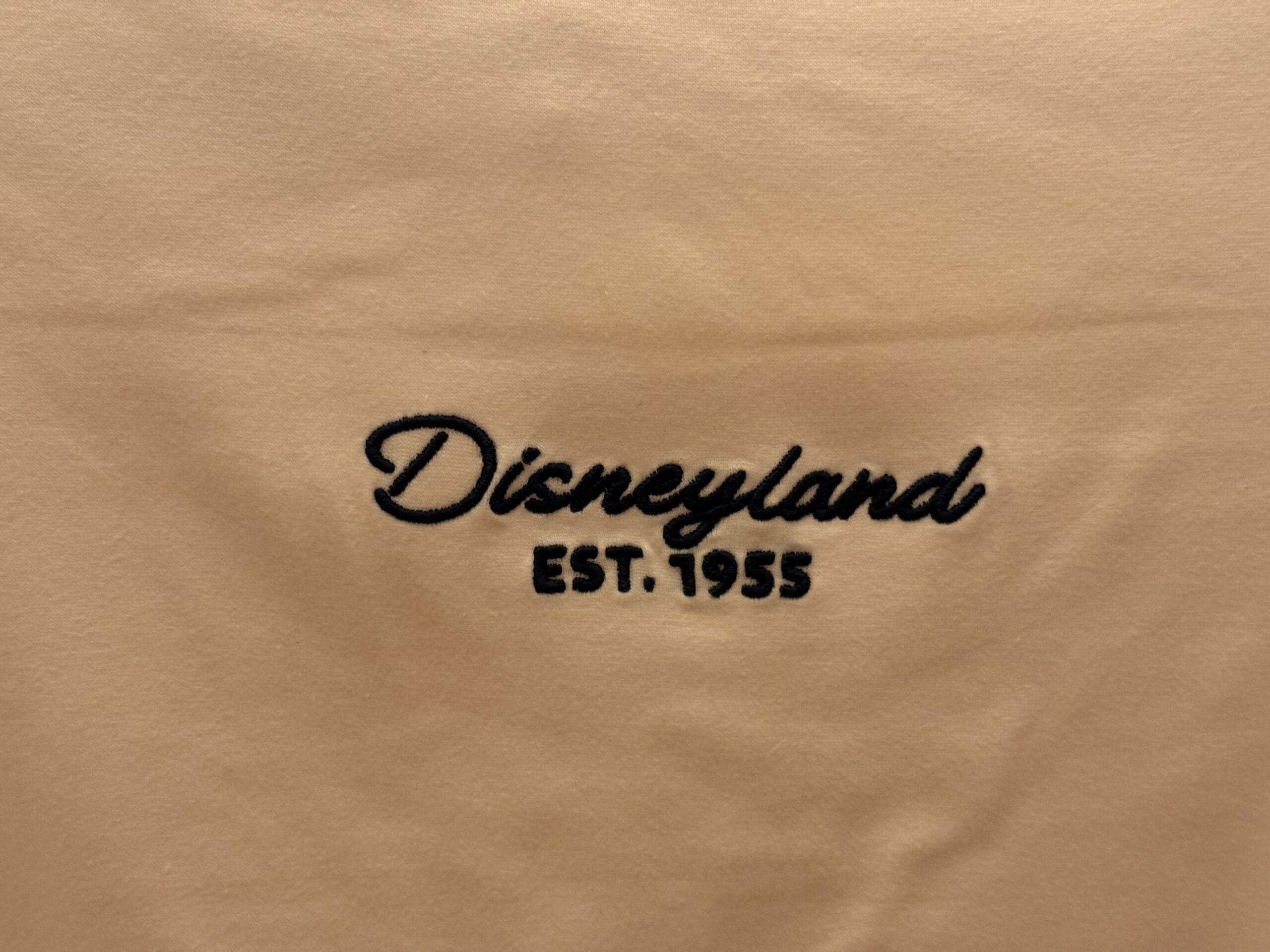 Beige fabric with "Disneyland EST. 1955" embroidered in black script and block letters, referencing the original Anaheim park.