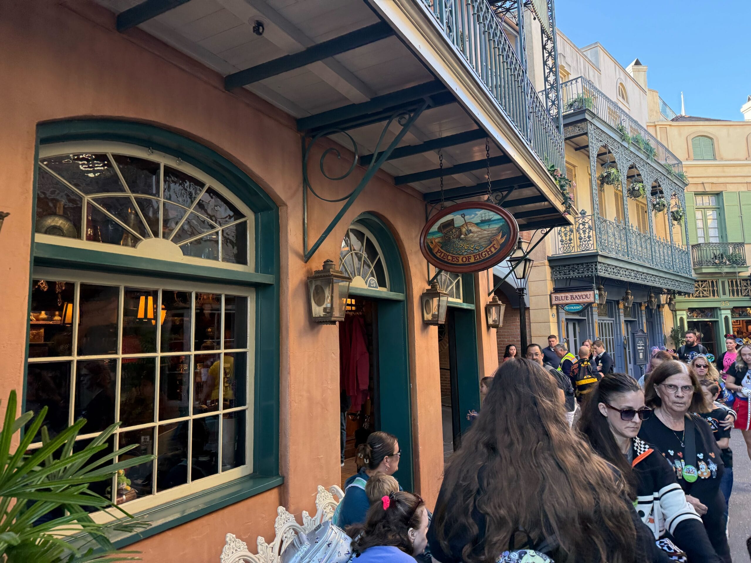 Guests stroll past a peach-colored building with green trim in a bustling shopping area, reminiscent of Main Street U.S.A. at Disney parks.