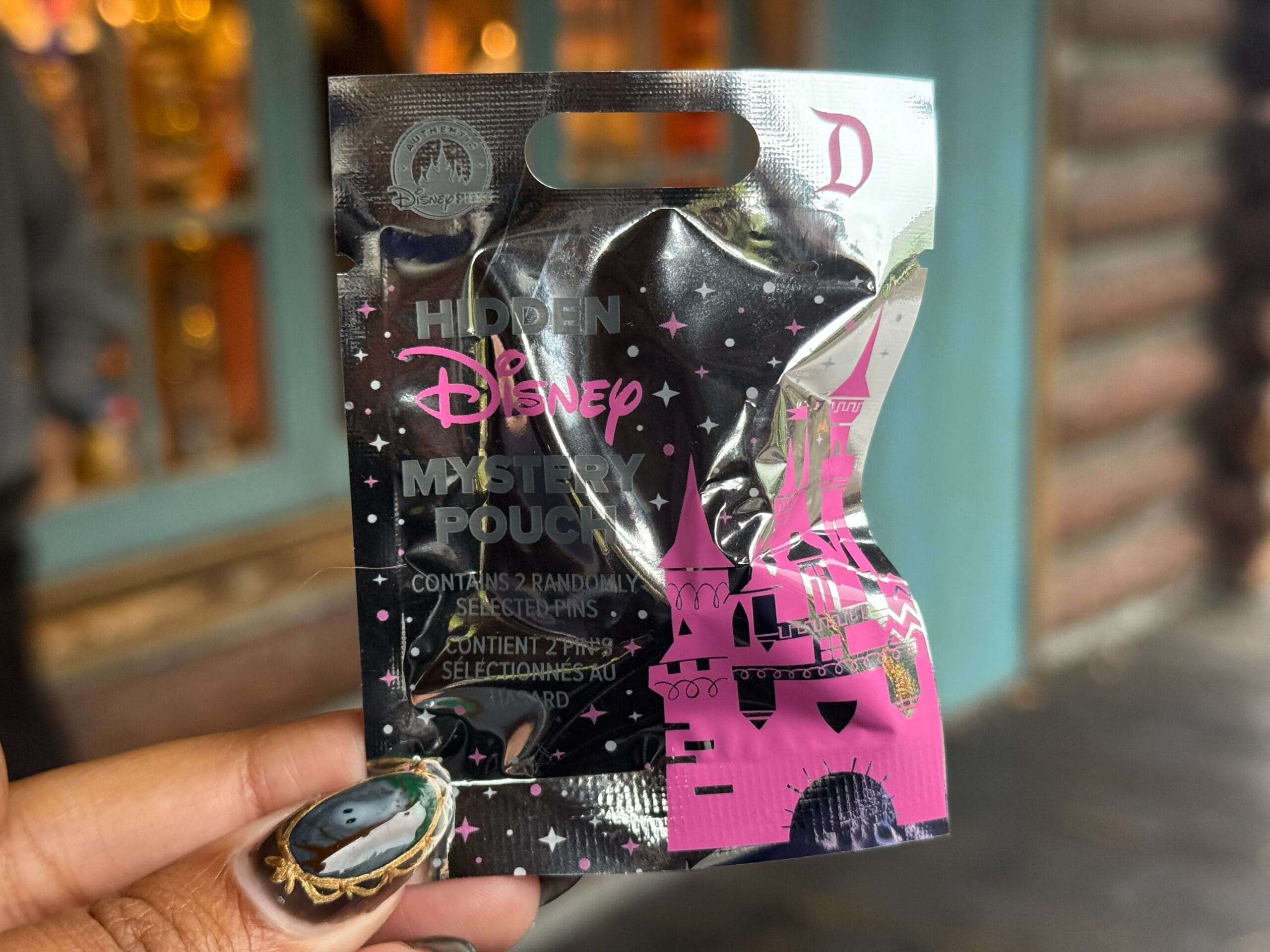 A hand holds a shiny foil Disneyland Hidden Disney Pin Mystery Pouch featuring pink Sleeping Beauty Castle art.