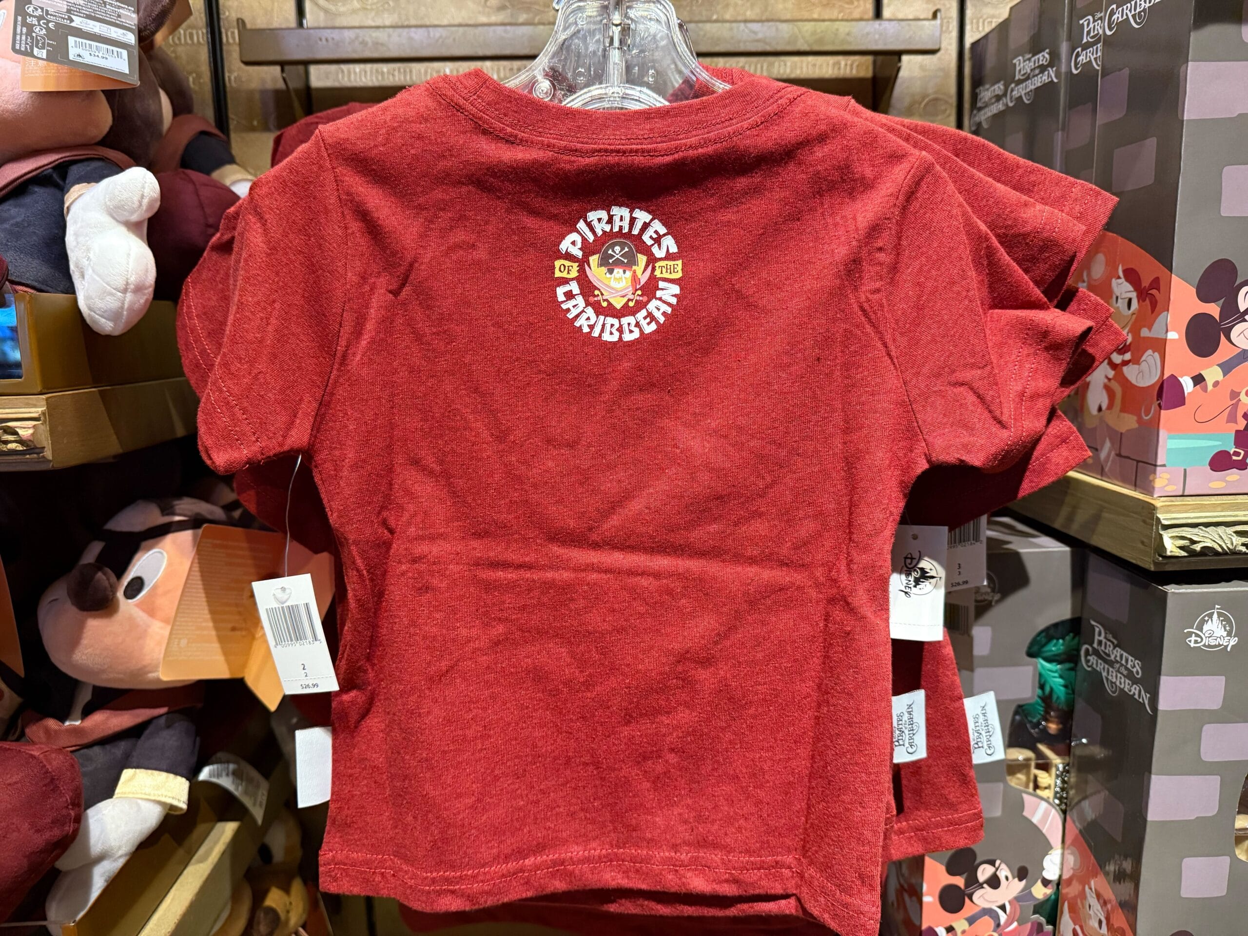 A red Pirates of the Caribbean t-shirt in a Disney park shop, surrounded by plush toys, capturing classic theme park merch.
